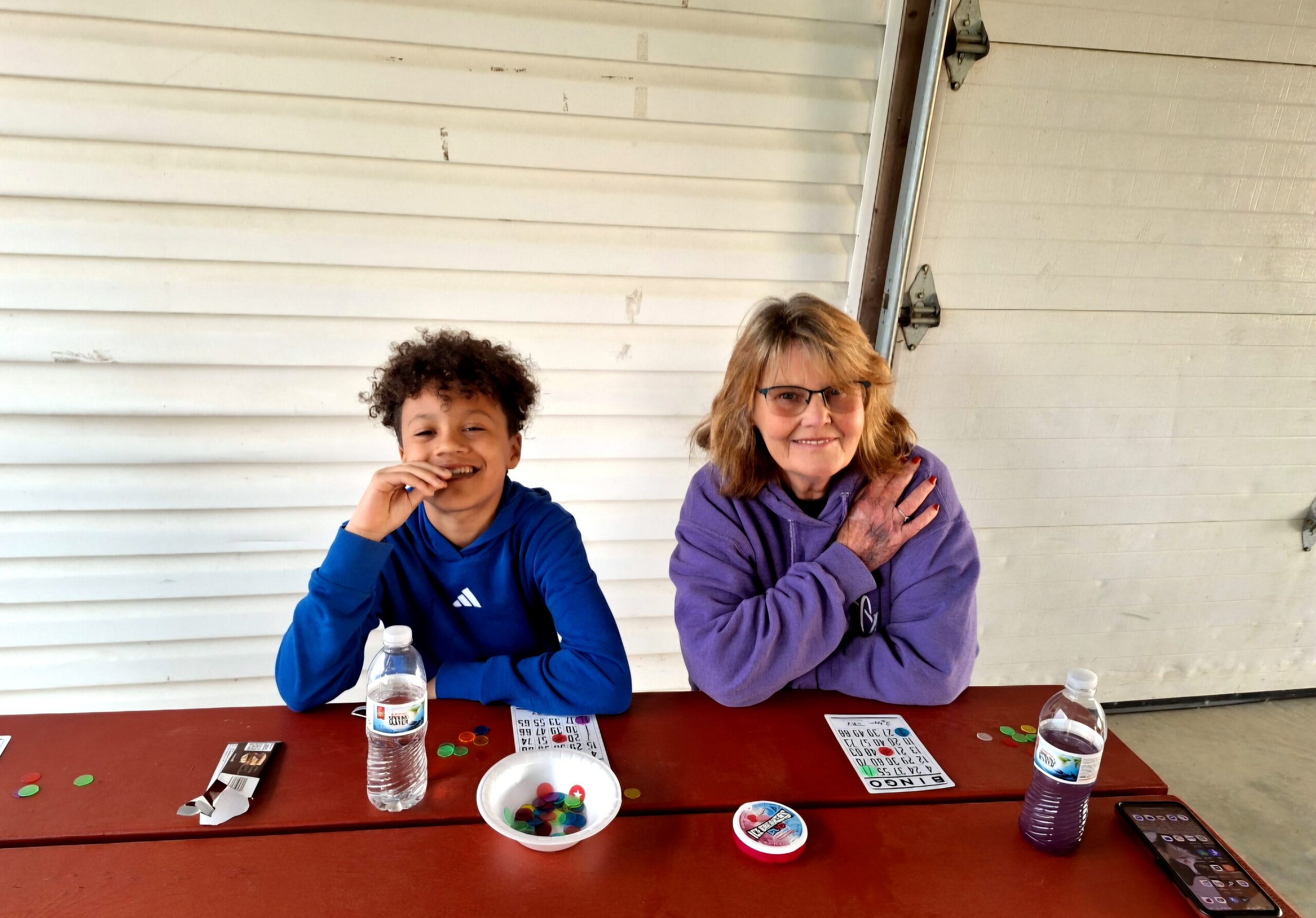 Two people sit at a red table playing a game with bingo cards, colorful chips, and water bottles visible on the table.