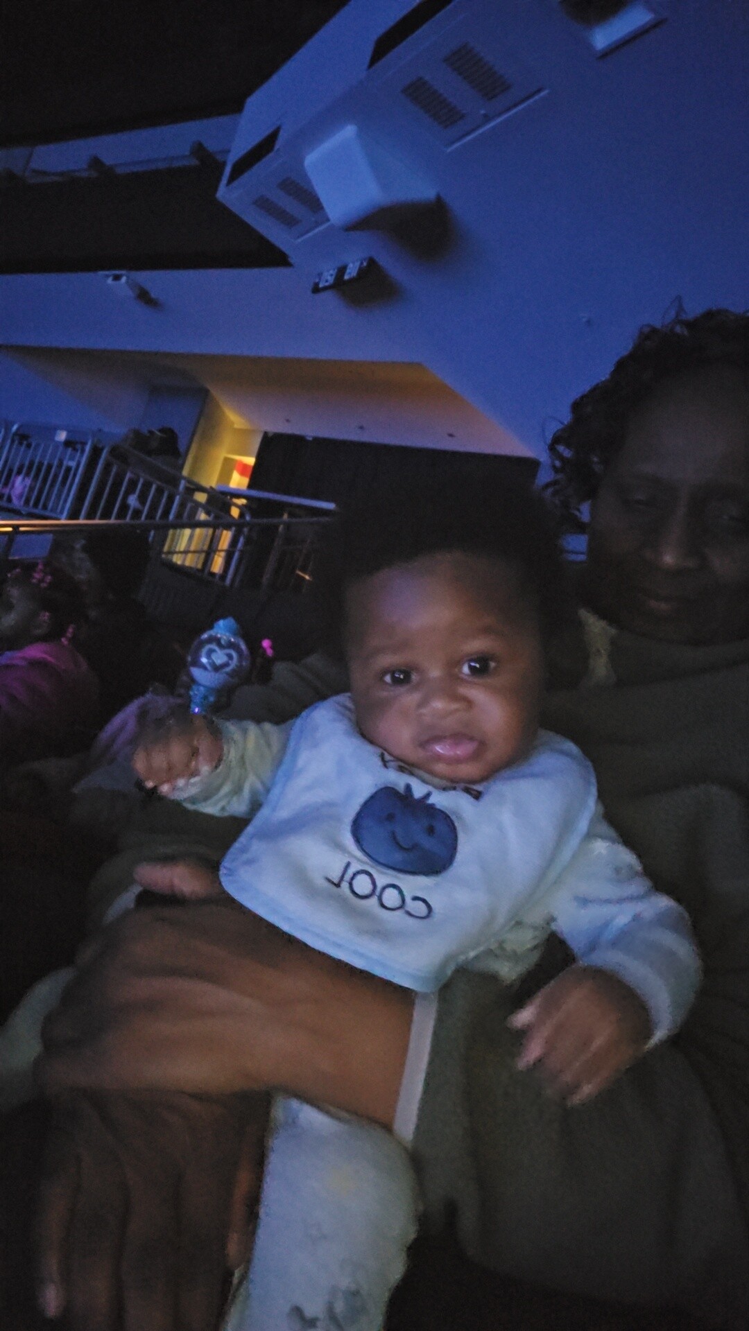Close-up of a baby in a light blue bib being held by an adult in a dim blue-lit room.