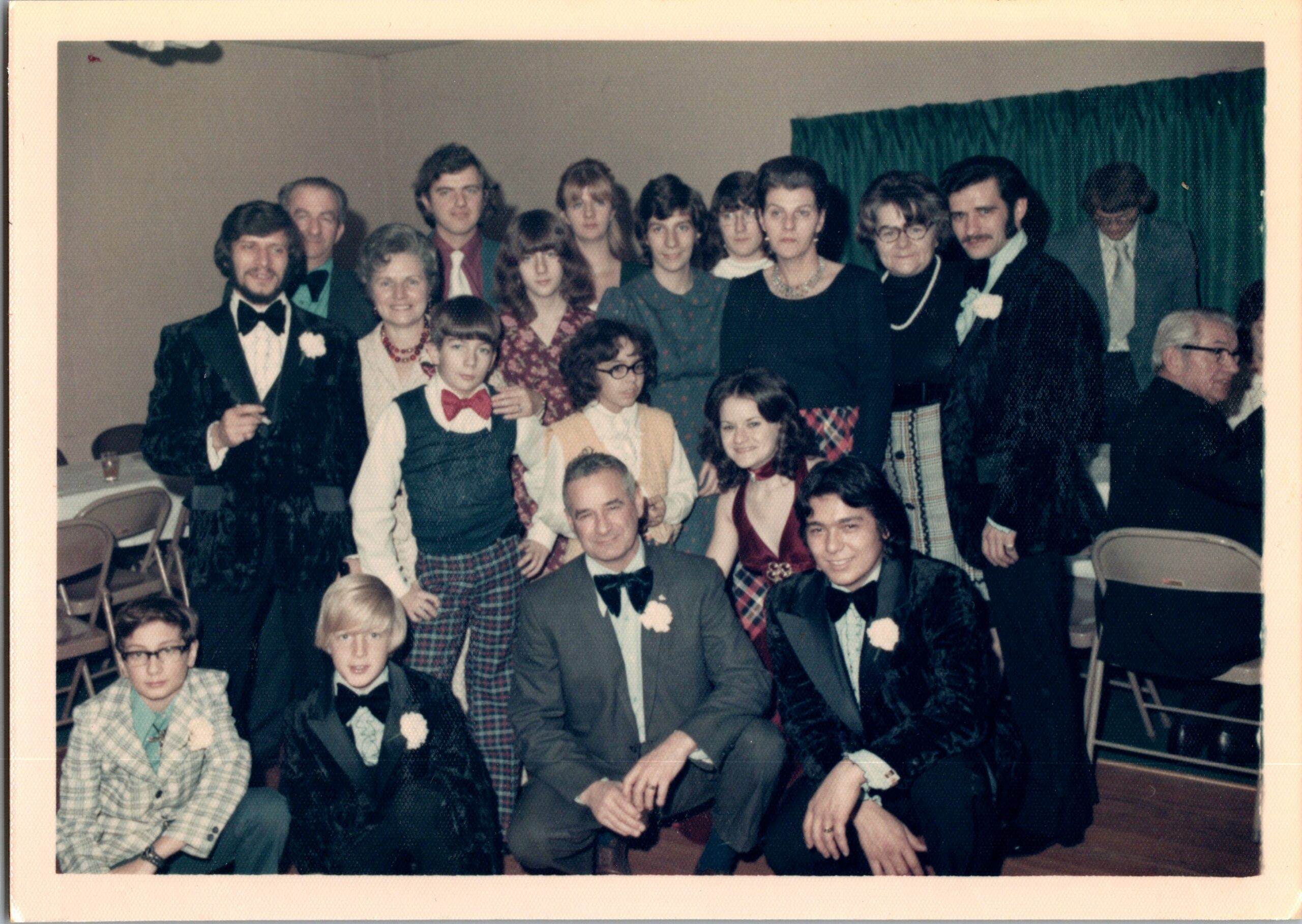 Group of people in formal attire posing for a photo at a party; men in tuxedos, women in dresses, indoor event space with chairs nearby.