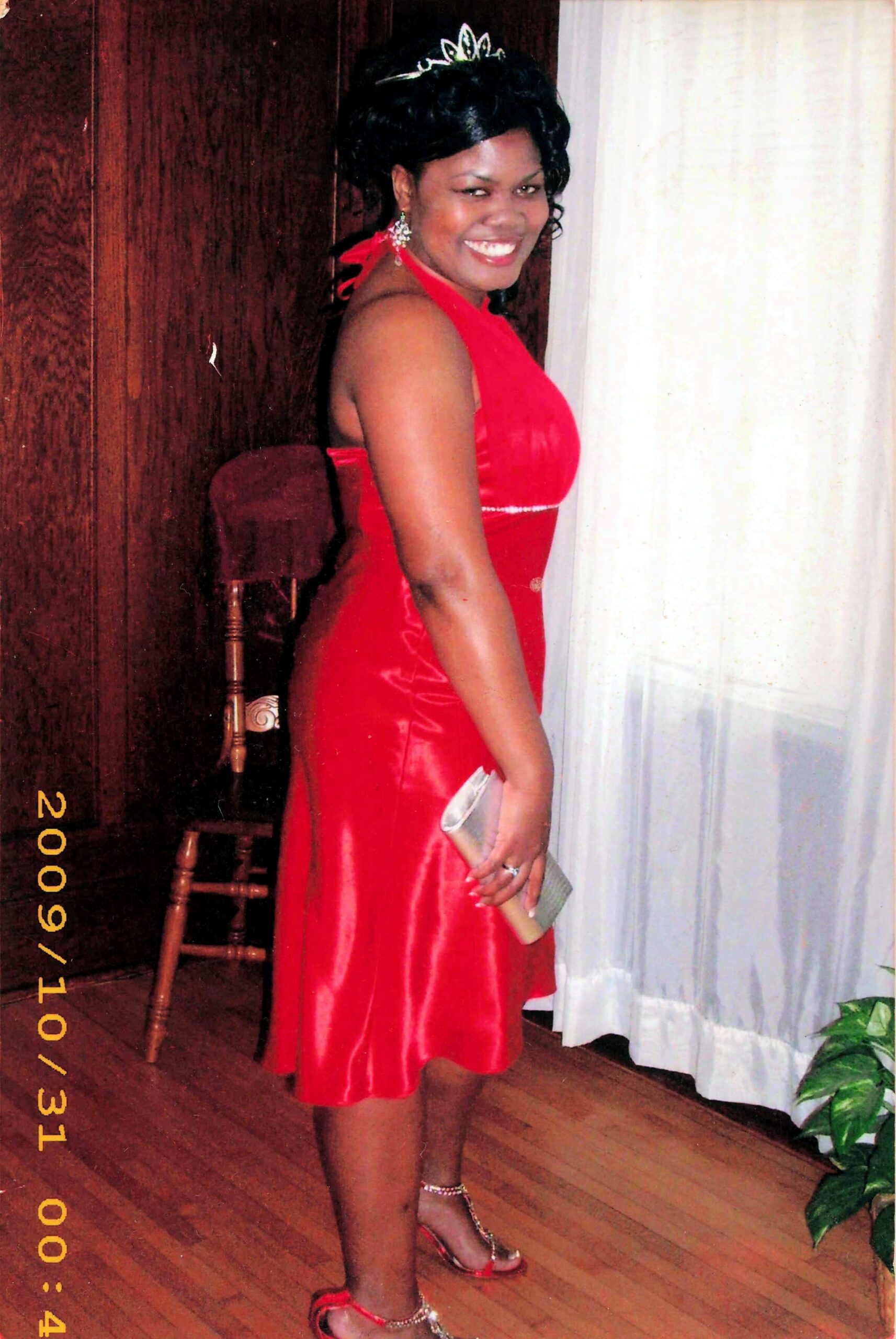 Smiling woman in a red satin dress with a tiara, holding a clutch, standing indoors by a white curtain.
