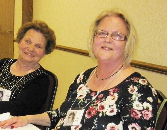 Two smiling women seated at a table in a meeting room, wearing name badges and lanyards.”] ,