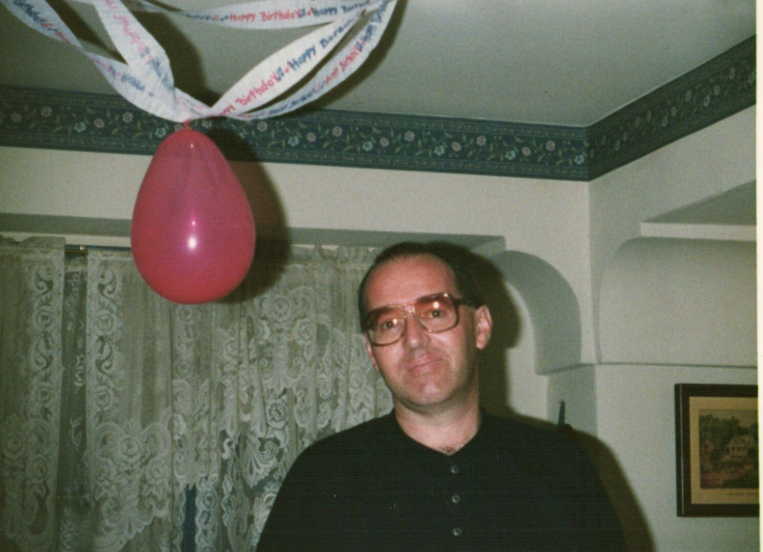 Man with glasses at a home birthday party, pink balloon tied to ceiling streamers overhead.