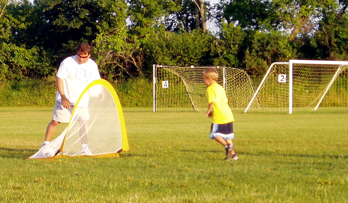 Adult in white helps a child kick a ball into a yellow-framed mini goal on a sunny grassy field.