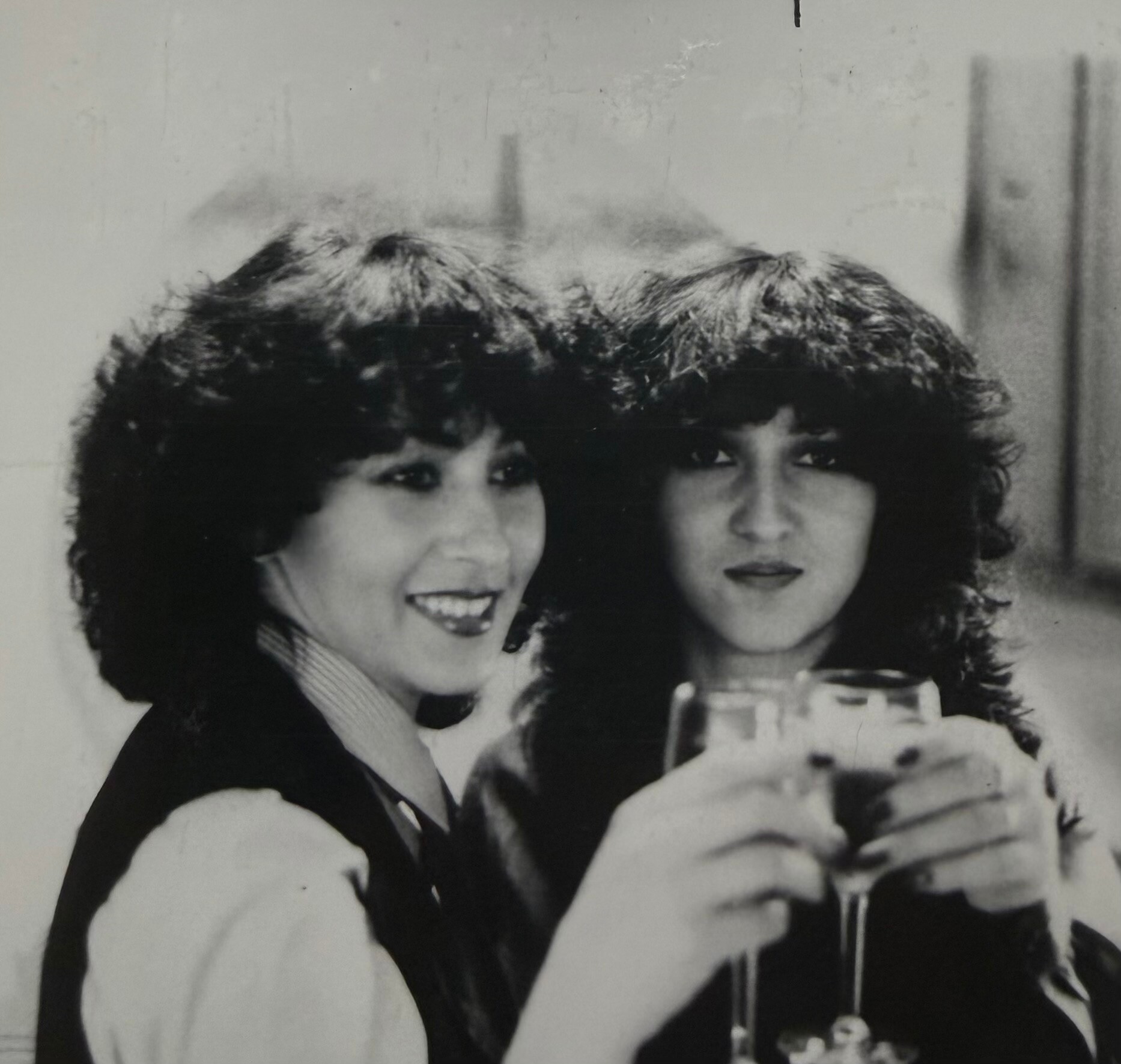 Two women in formal attire smiling at the camera, each holding a stemmed glass in a black-and-white photo.