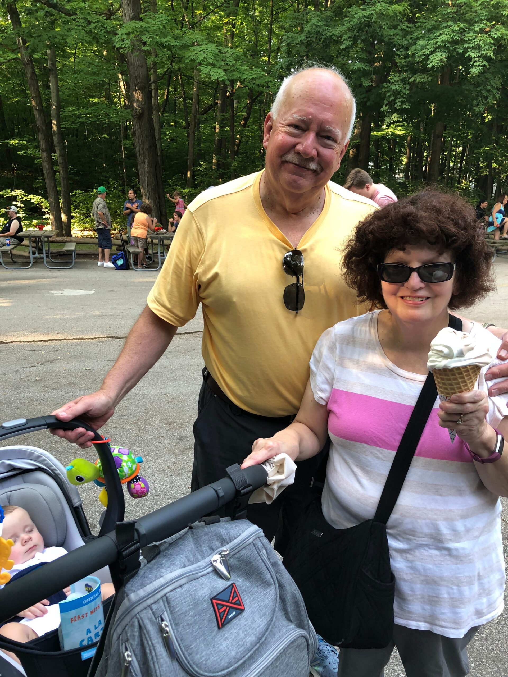 Smiling older man in a yellow shirt stands beside a woman with sunglasses in a park, pushing a baby stroller with a sleeping infant.