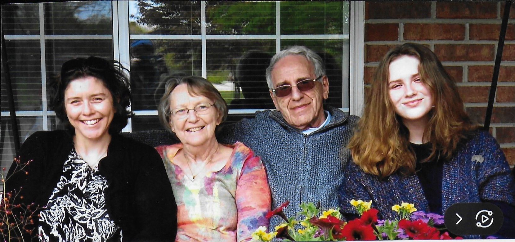 A smiling family of four sits on a porch with flowers in front, in front of a brick wall and window.