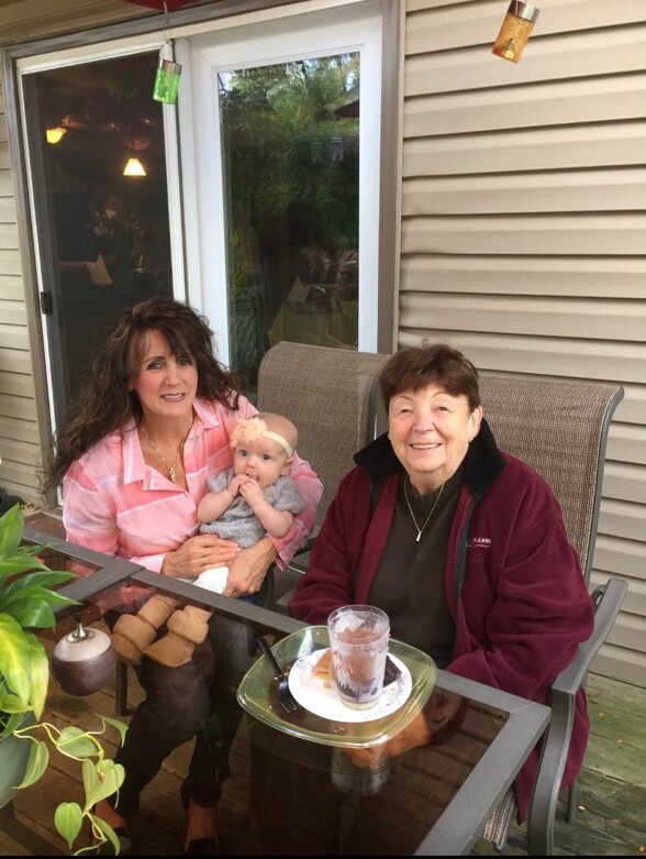 Three generations sit on a porch: a woman in pink holding a baby, next to an elderly woman in a maroon jacket smiling outdoors.