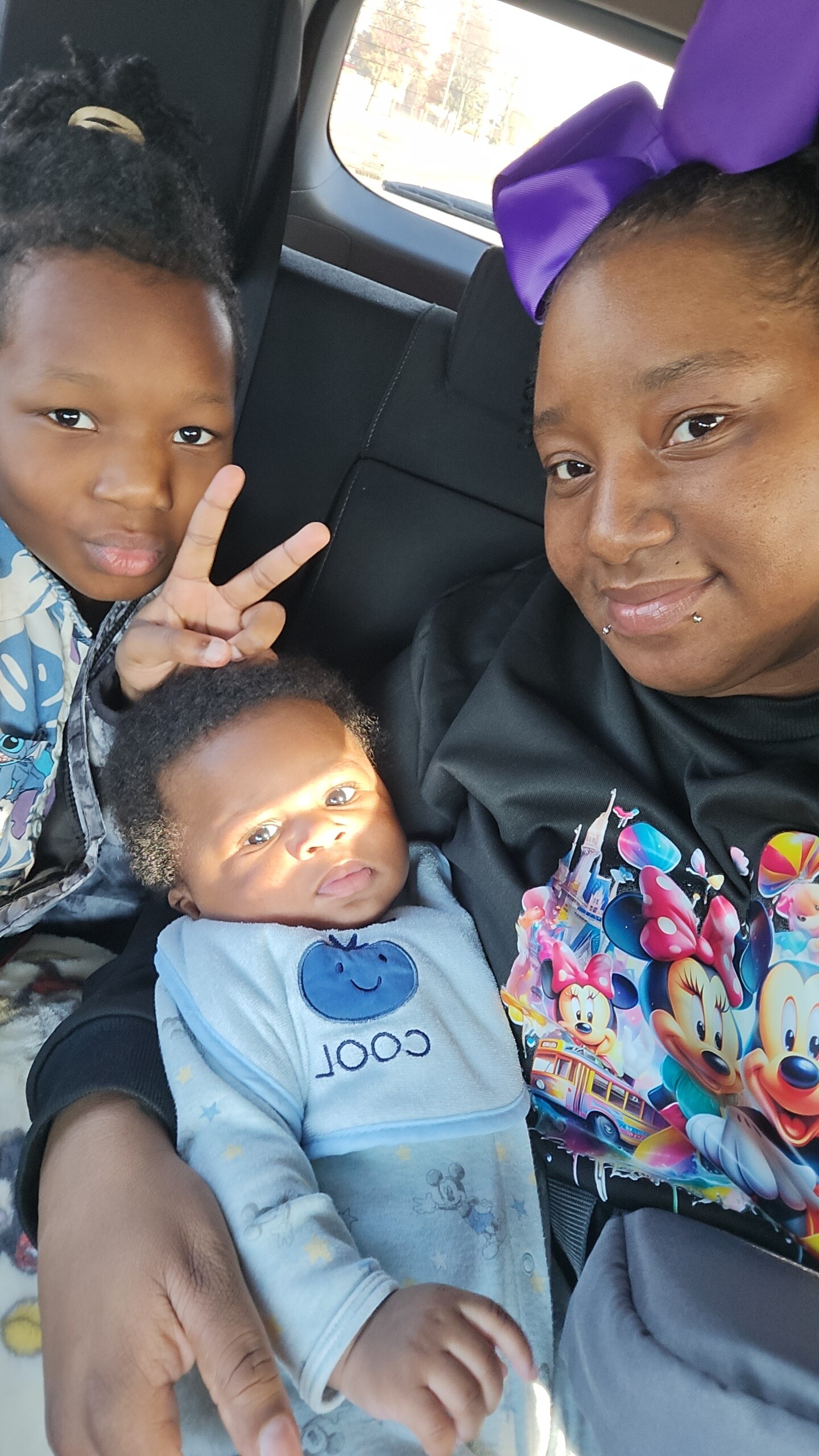 Woman with two children in a car: older girl making a peace sign, baby in a blue onesie looks at the camera next to her.