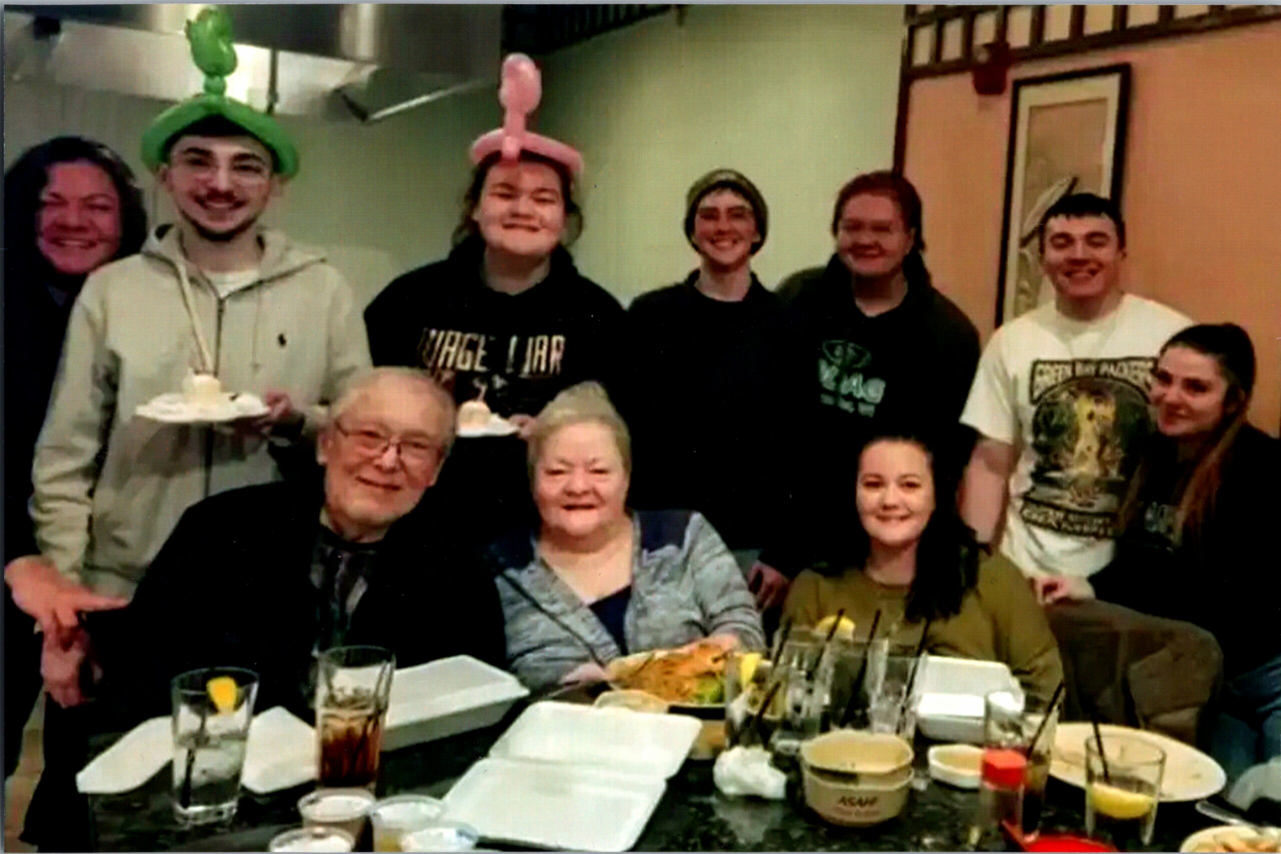 Group of people gathered around a dining table at a celebration, some wearing balloon hats, smiling. (informational)