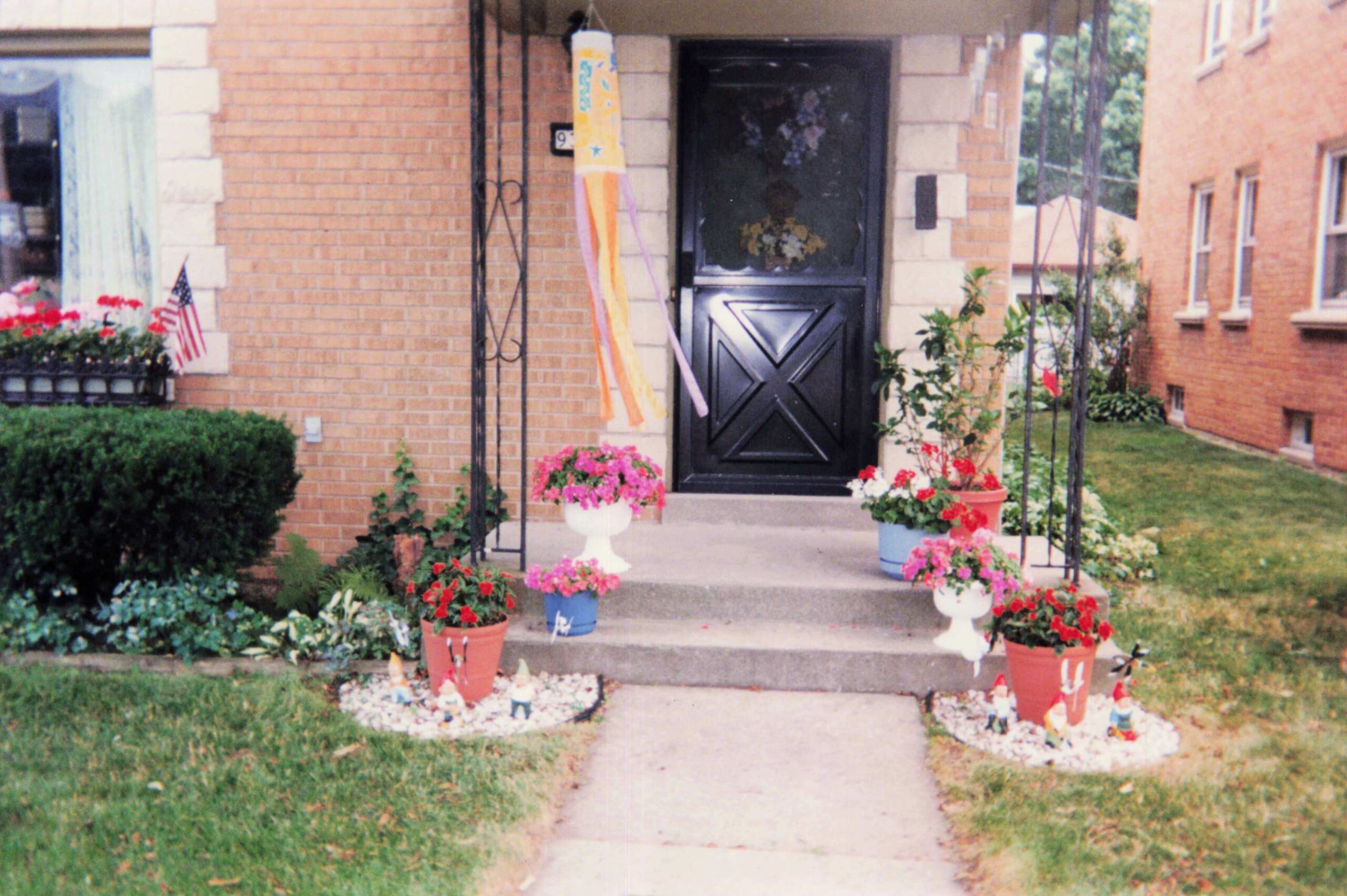 Front porch of a brick house with a black door, colorful streamers hanging near the entrance, and potted flowers along the concrete steps. A small, decorative flower bed with white rocks sits at the walkway, and a green lawn surrounds the area.
