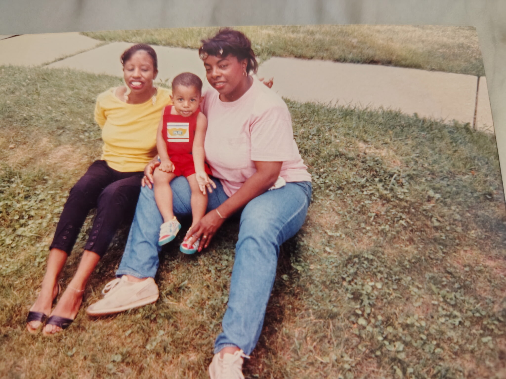 Three people sit together on a grassy area outdoors: a woman in yellow, a small child in red overalls, and a woman in a light pink shirt, smiling at the camera.