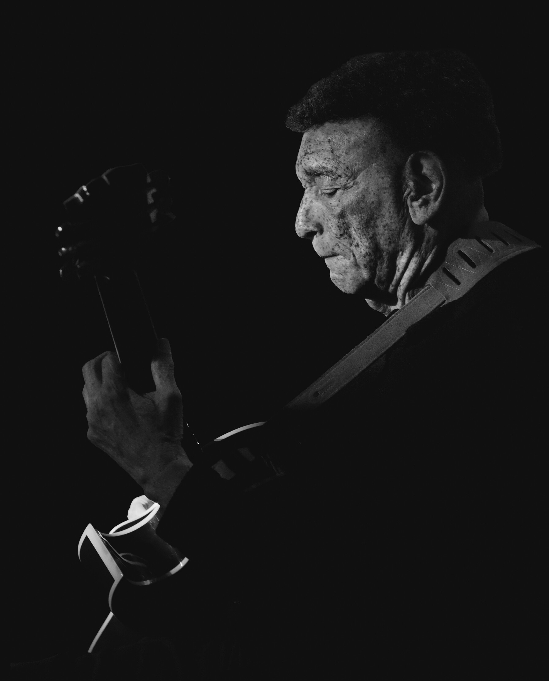 Side-profile of a man with a guitar in a dark setting, concentrating on playing the instrument (black-and-white photo).