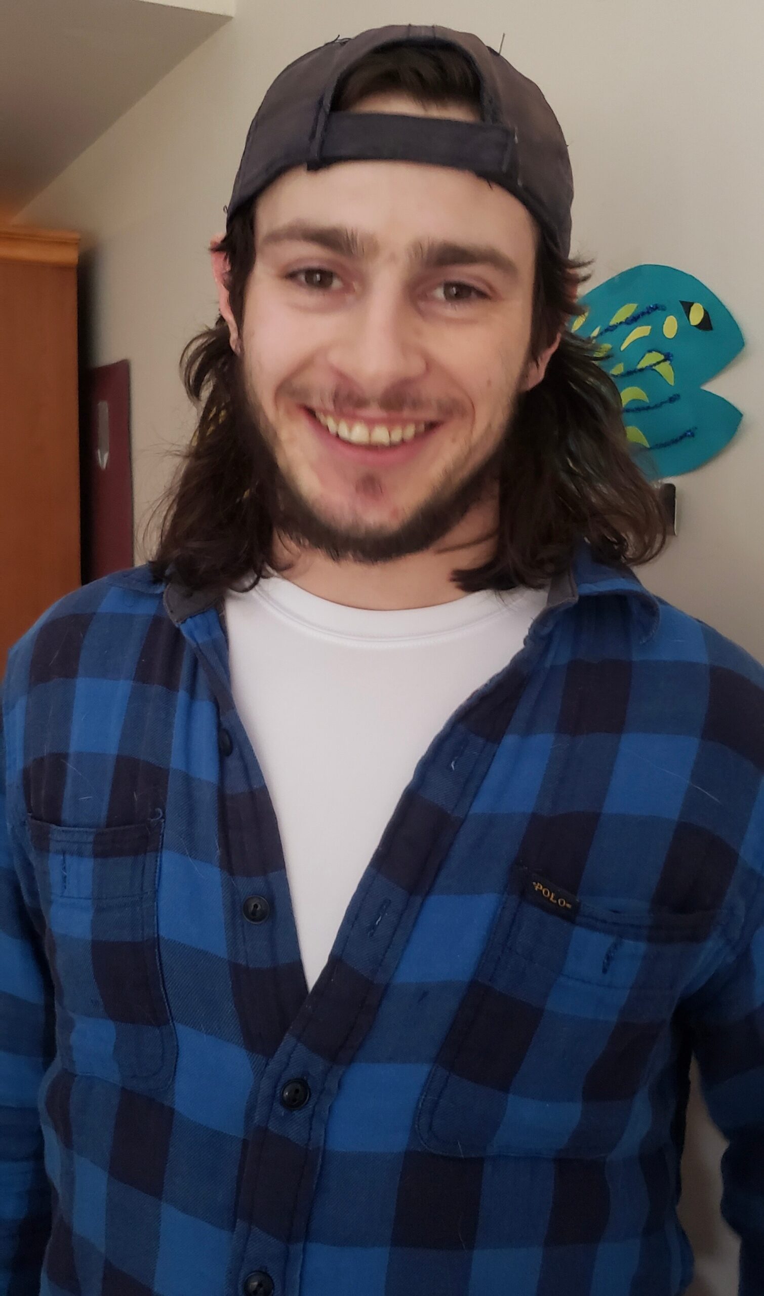 Young man with a backward cap and long hair, wearing a blue plaid shirt, smiling at the camera on screen life photo-style portrait.