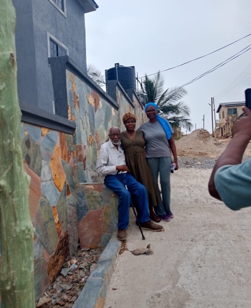 Three people posing for a photo along a mosaic-tiled wall; an elderly man with a cane sits while two women stand beside him under a blue sky, a photographer capturing the moment on the right.