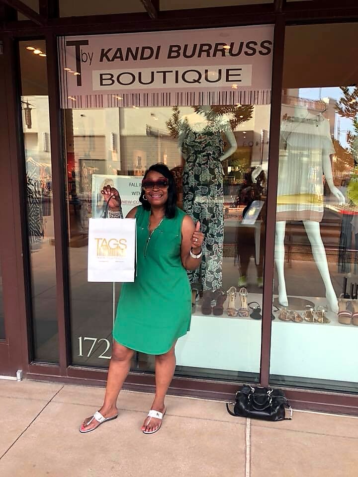 Woman in a green sleeveless dress stands outside Kandi Burruss Boutique, holding a white shopping bag and smiling.