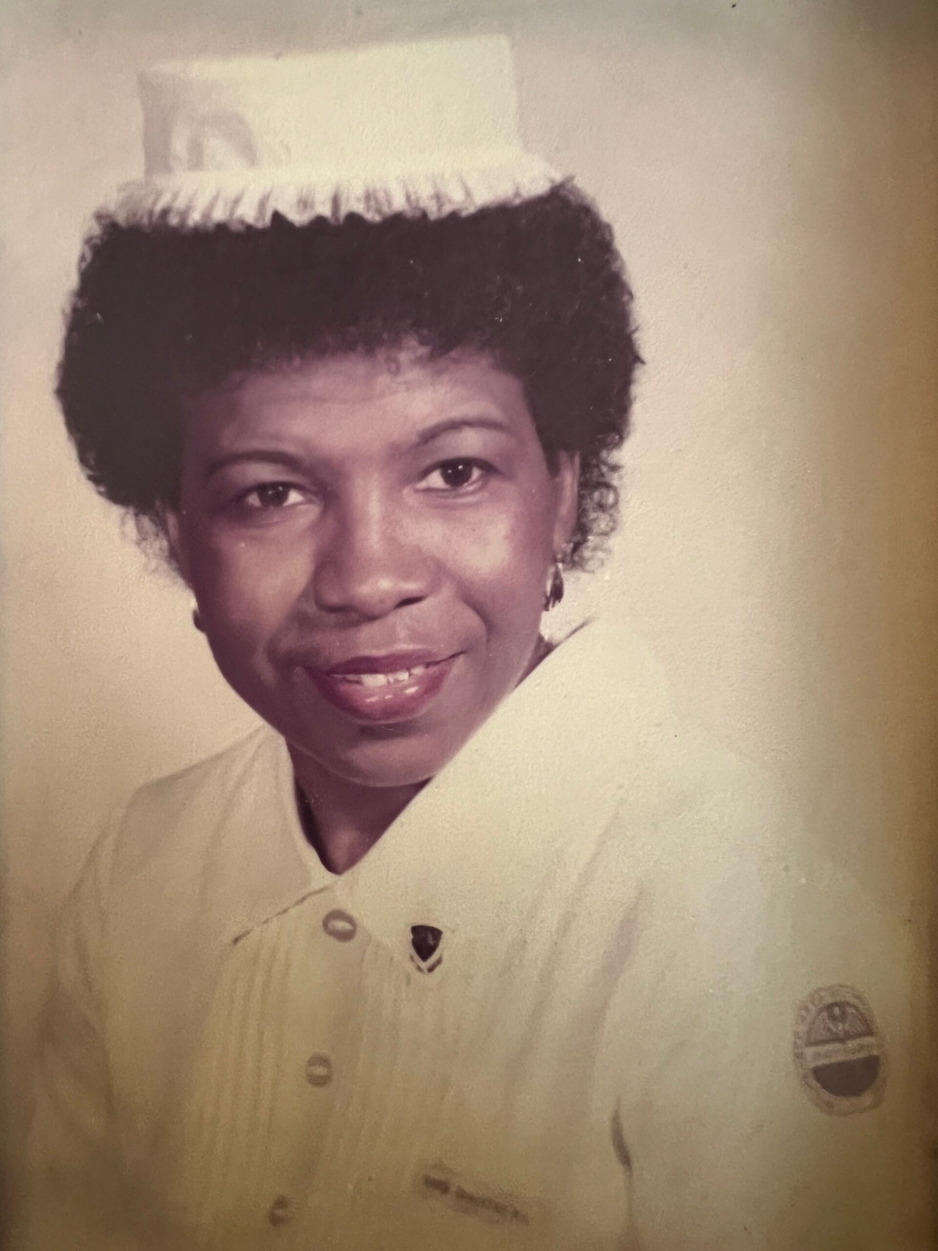 Portrait of a woman in a vintage white nurse uniform with a small white cap, smiling at the camera.