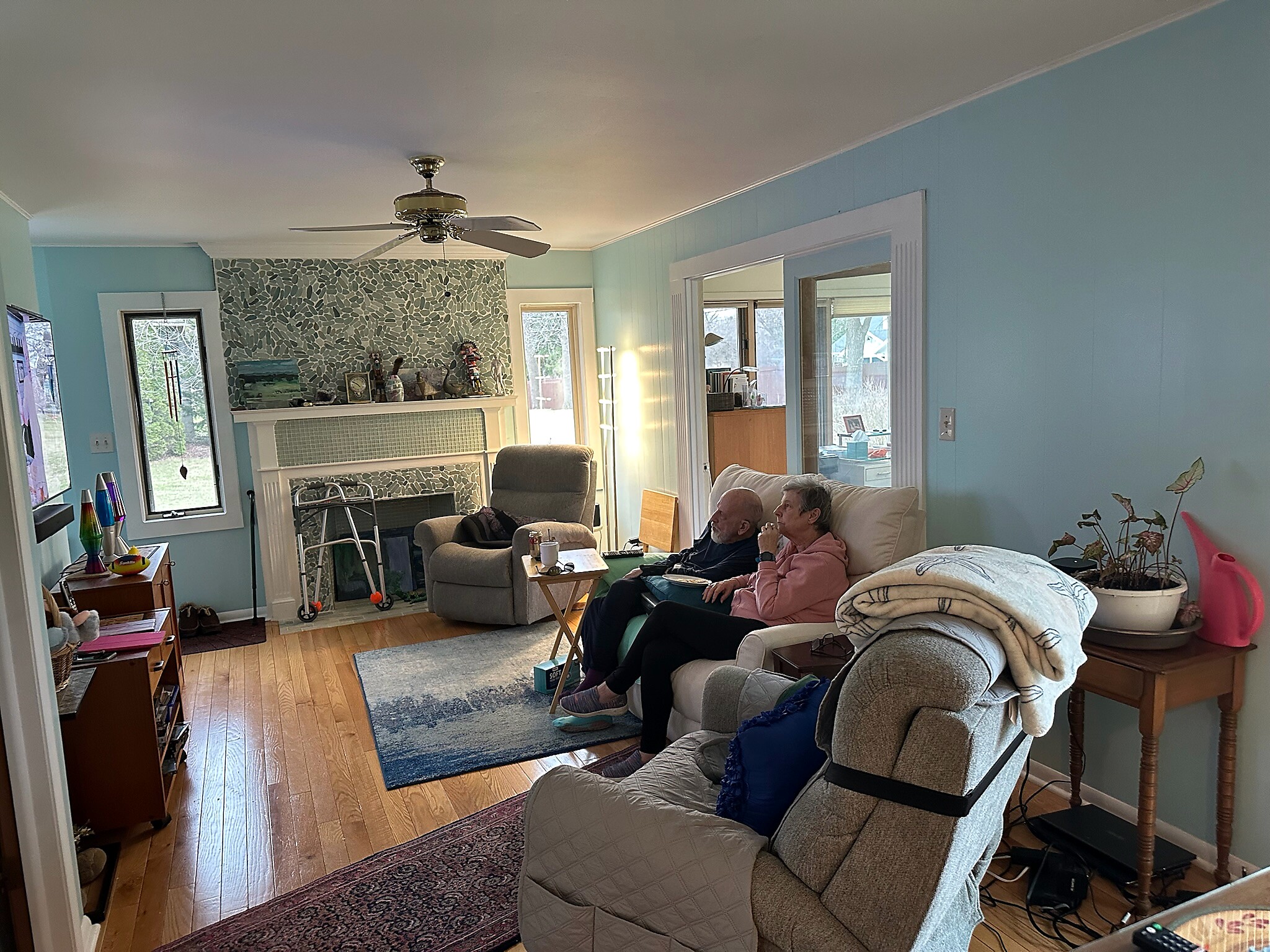Living room with light blue walls, mosaic fireplace, and ceiling fan; two people sit on a sofa and chair.