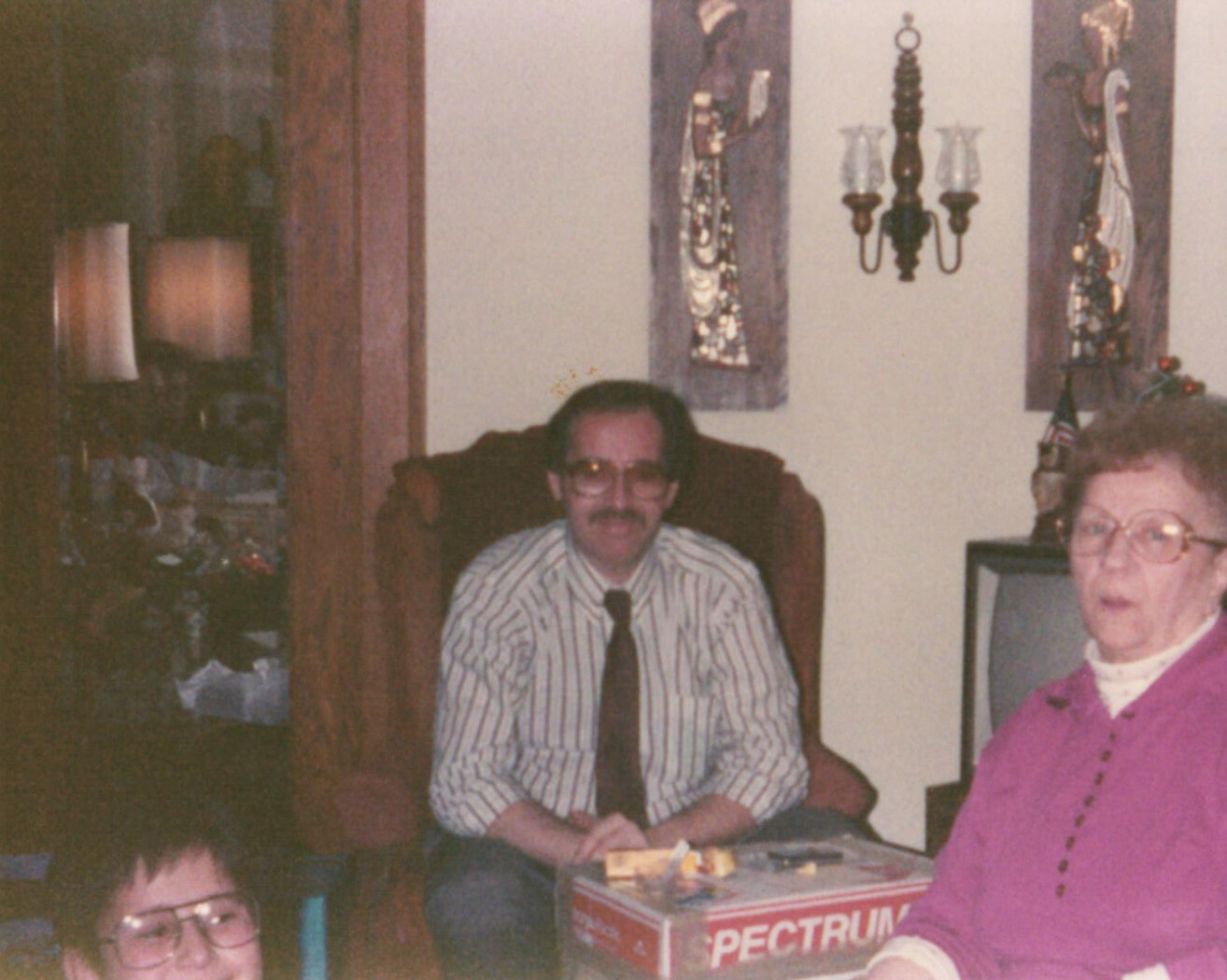 Man wearing a striped shirt and tie sits in a brown armchair in a living room, with a woman in a pink sweater nearby at a table with a Spectrum box in view.