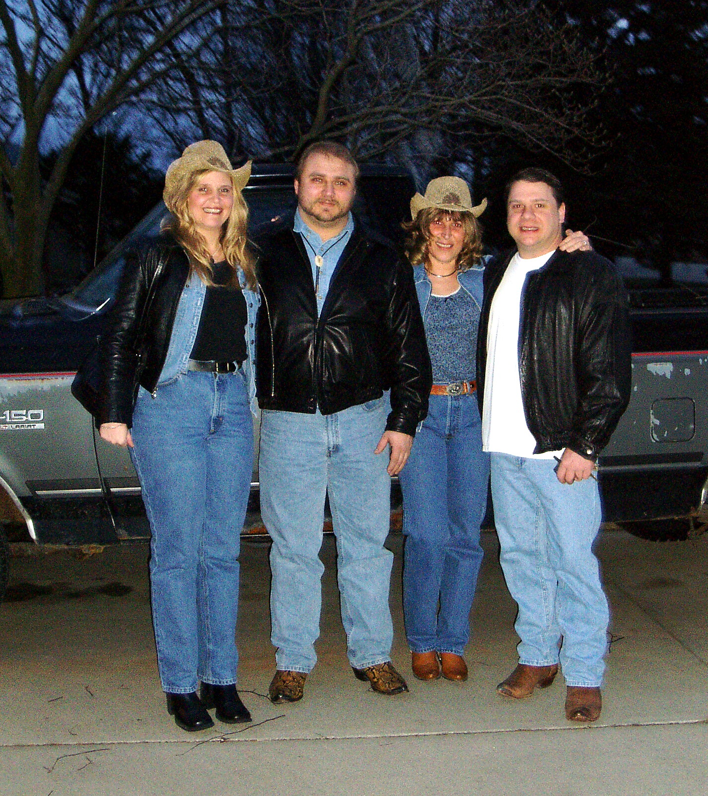 Four adults in denim and leather jackets posing together outdoors at night, two wearing cowboy hats.