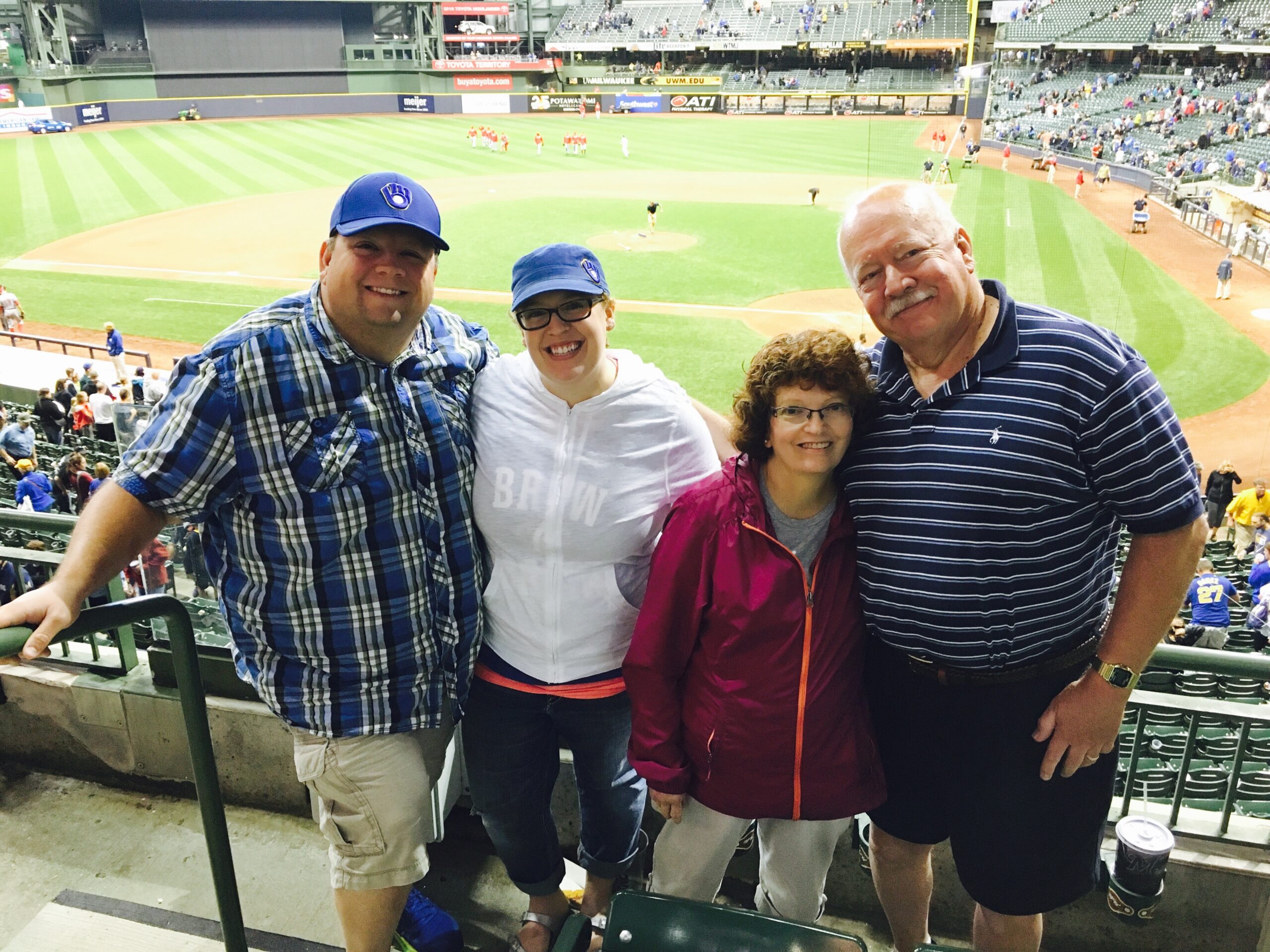 Four smiling fans stand at a baseball stadium railing, posing for a photo with the field and players in the background.