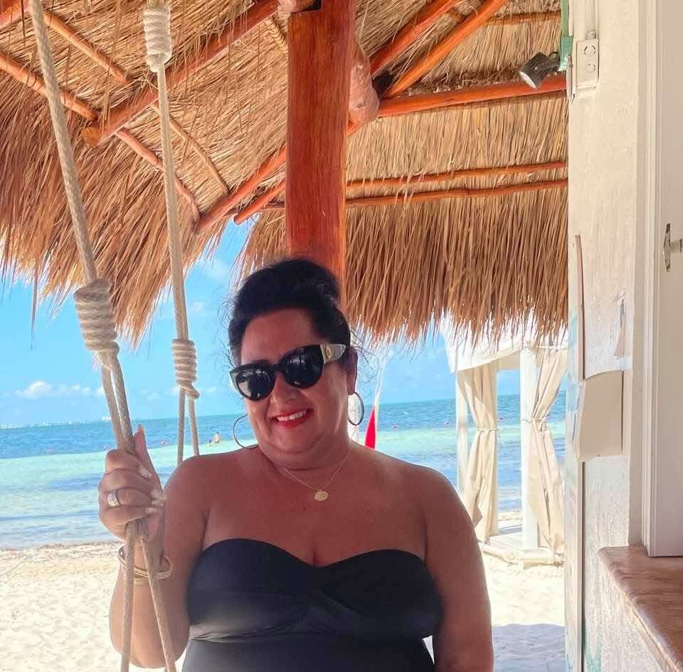 Smiling woman in a black strapless swimsuit and sunglasses stands on a sunny beach, holding a rope swing under a thatched cabana with the sea in the background.