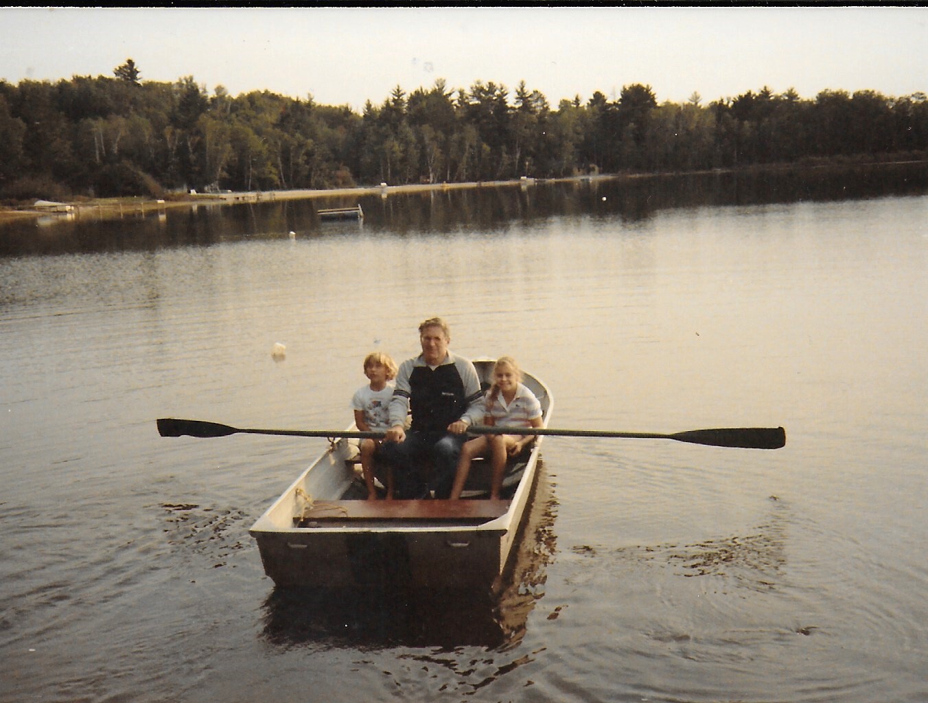 Family of three in a small rowboat on a calm lake with a forested shore in the background, paddling together.
