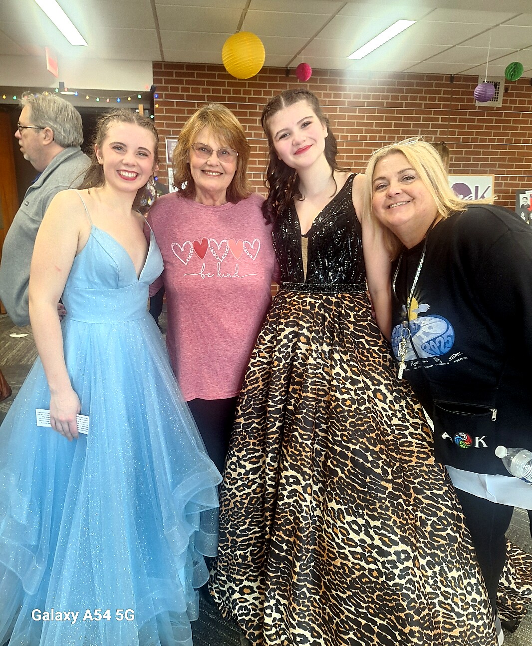 Group photo at a party: four smiling people standing indoors, two girls in formal dresses (blue ball gown and leopard-print gown) with two adults nearby, brick wall and party decorations in the background.