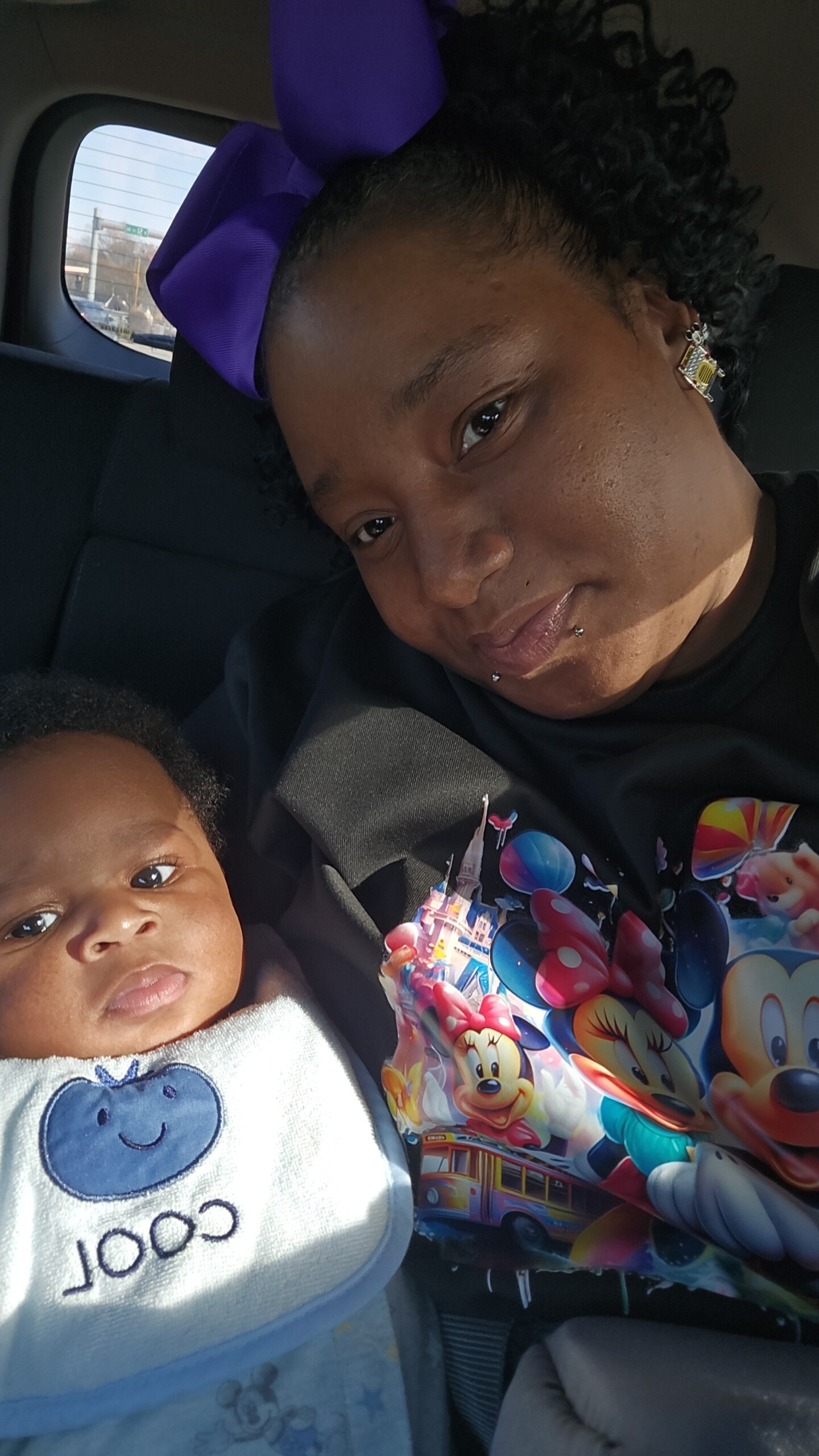 Woman and baby in a car seat together; the woman wears a purple bow and colorful shirt, smiling gently at the camera.