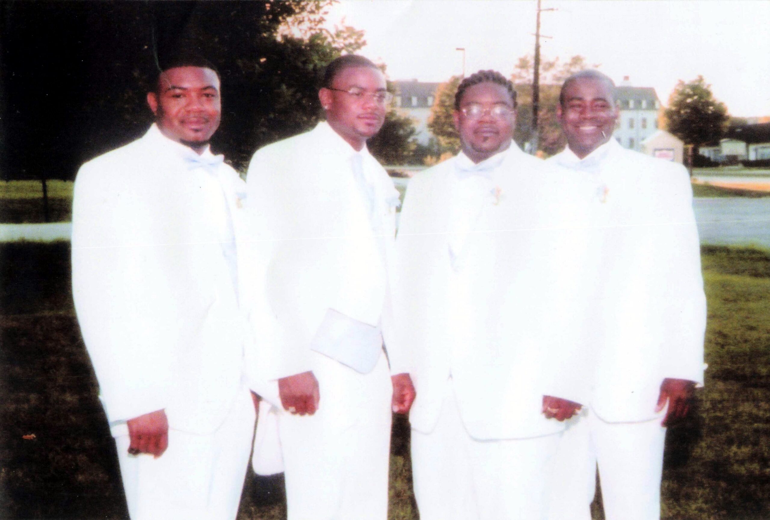 Four men in white tuxedos posing outdoors on a grassy area, smiling at the camera.