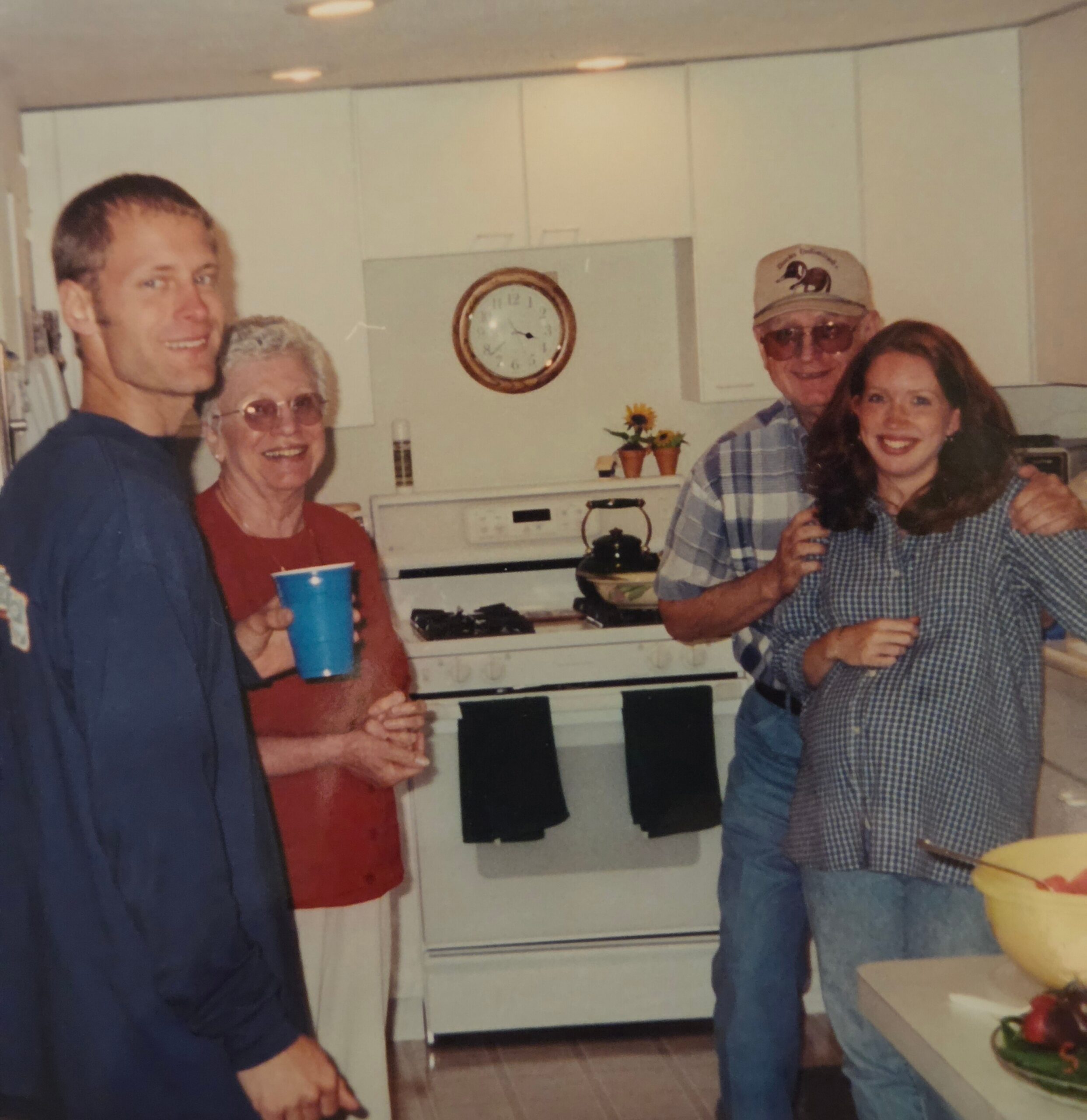 Four adults smiling in a home kitchen, posing for a photo with cabinets and a stove behind them.