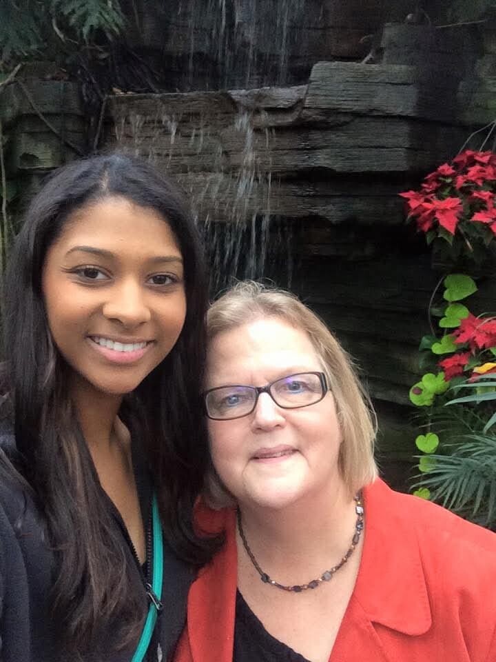 Two women pose for a selfie in front of a small waterfall with red flowers nearby, smiling at the camera.