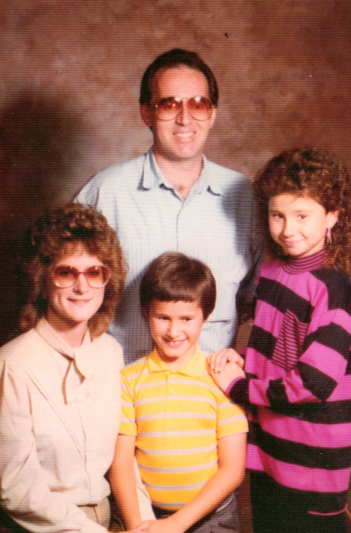 Smiling family portrait with a man in sunglasses standing behind three children in colorful clothing.