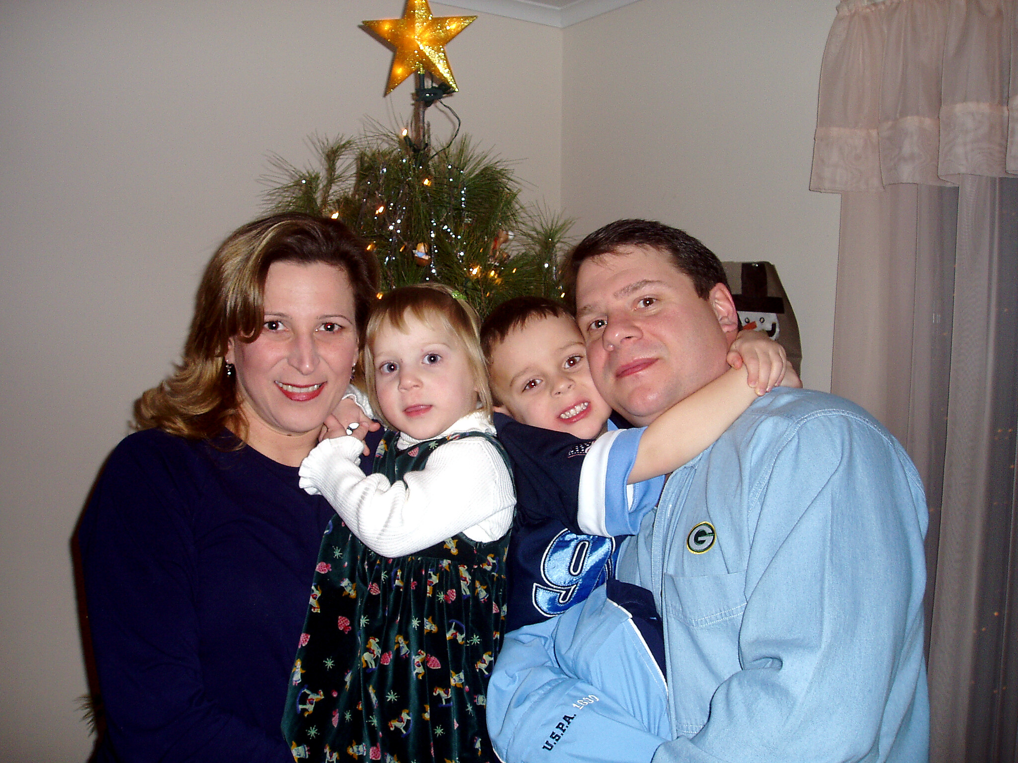 Family of five posing for a holiday photo in front of a decorated Christmas tree with a star top and lights.