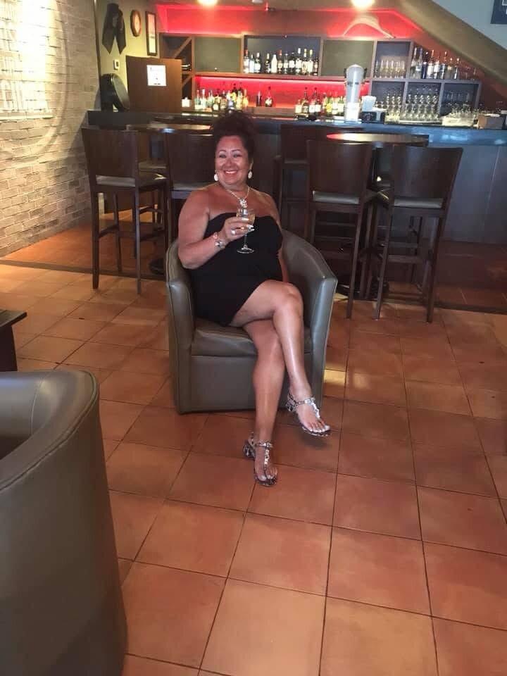 Smiling woman in a black strapless dress sits in a leather chair at a bar, holding a wine glass.
