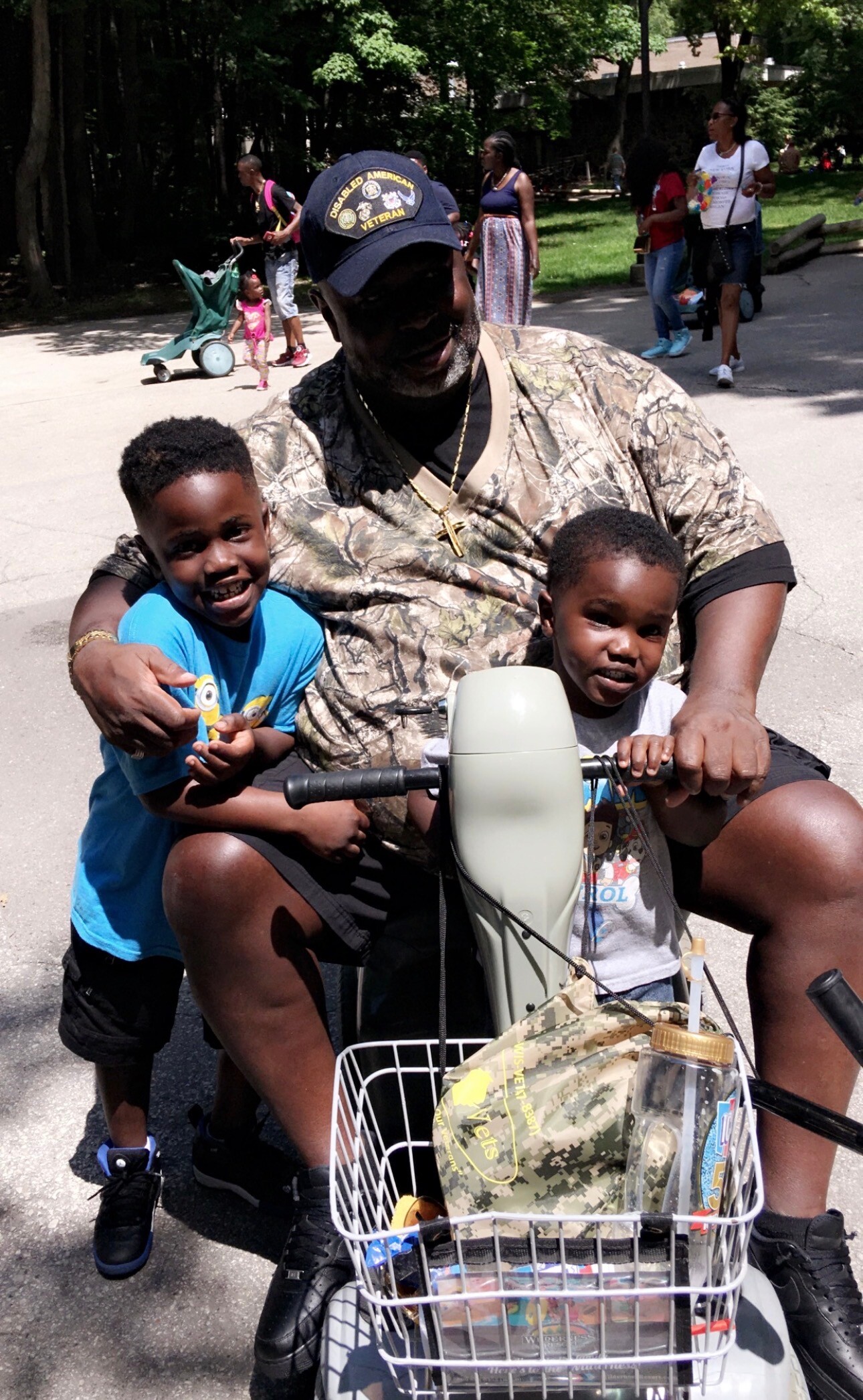 A man in camouflage shirt on a bike with two smiling children in front, riding in a park.