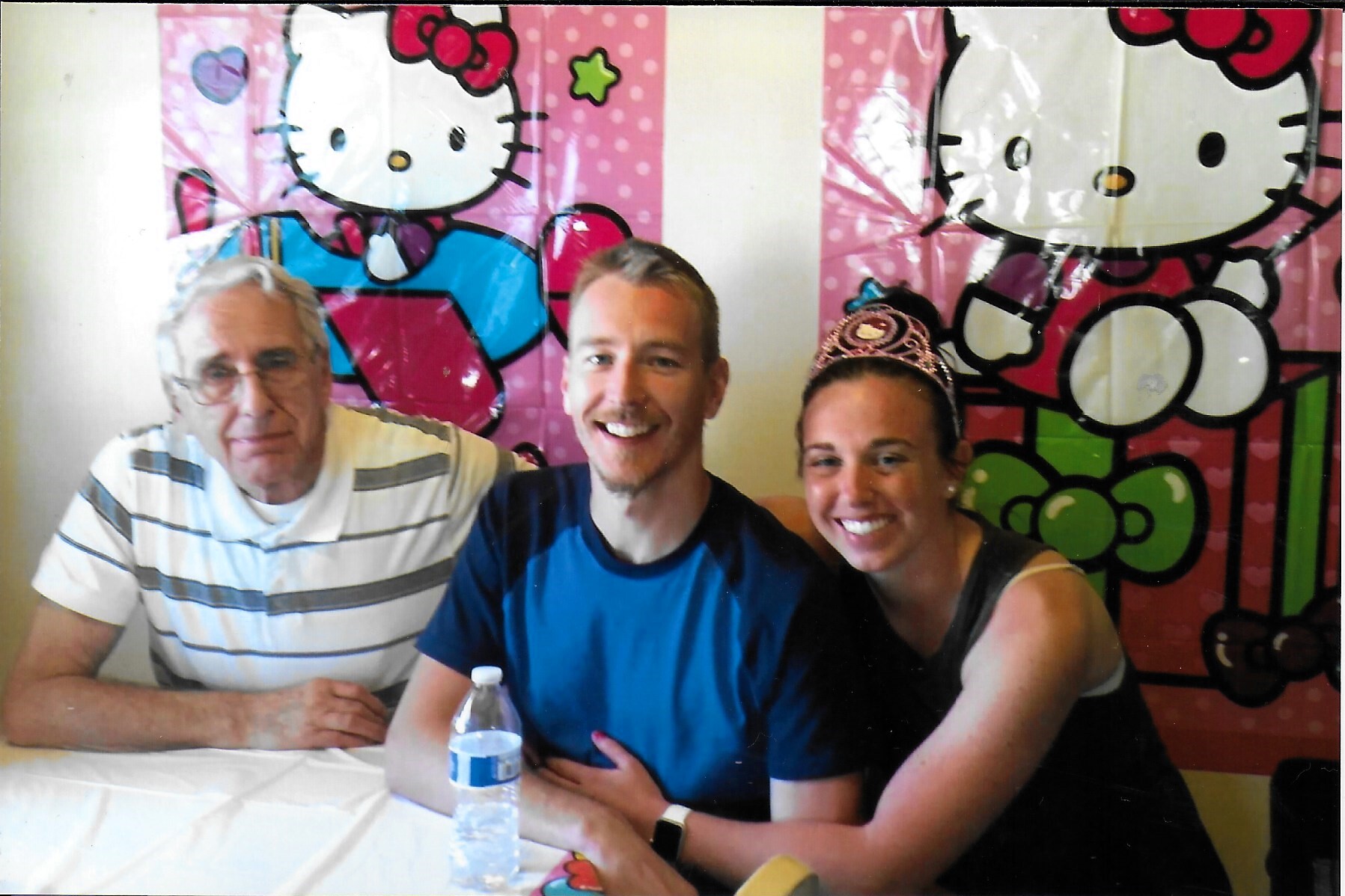 Three people sit at a table posing for a photo: a striped-shirt older man on the left, a smiling man in a blue shirt in the middle, and a woman wearing a tiara on the right, with a Hello Kitty mural in the background.