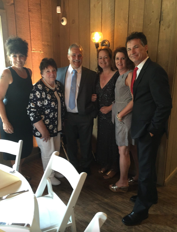 Six adults pose together for a group photo in a wood-paneled room, dressed in formal attire and smiling at the camera with dining tables and white chairs visible in the foreground.