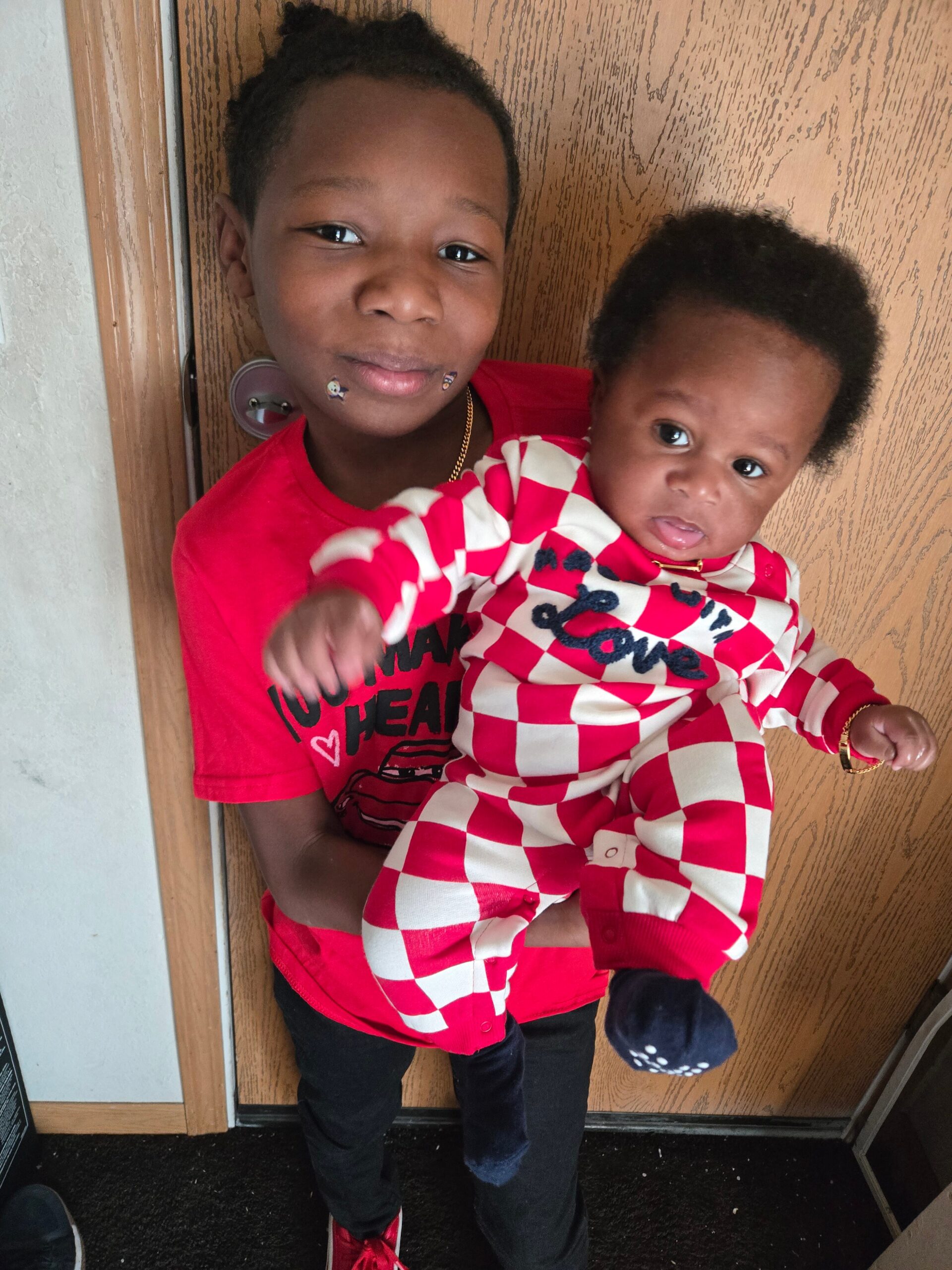 Older child in a red shirt holding a baby in a red-and-white checkered outfit by a wooden door.