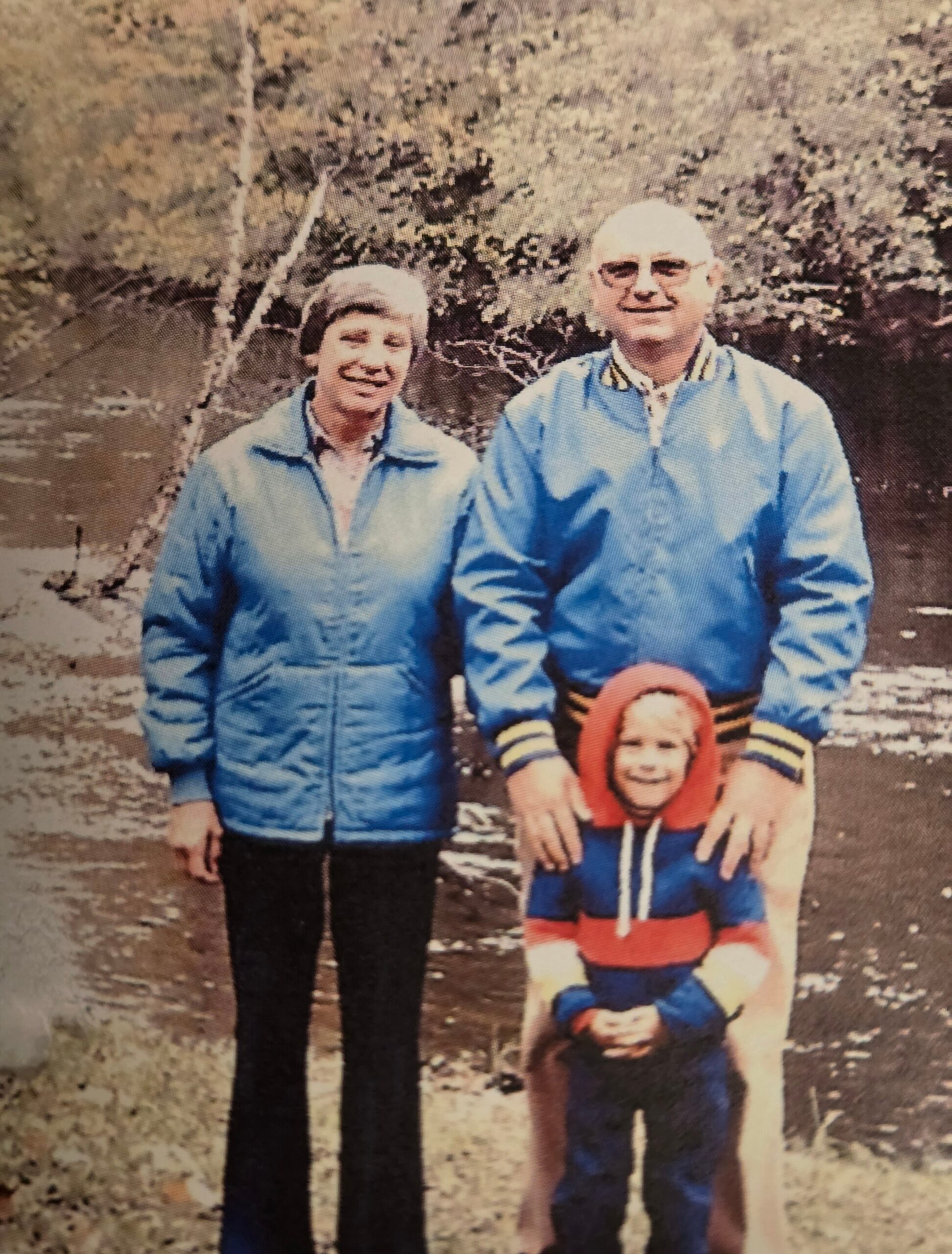 Family photo taken outdoors by a river; two adults in blue jackets stand with a child wearing an orange life vest in front, all smiling.