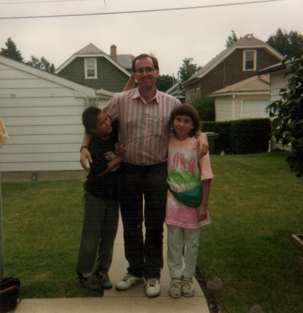 A man in a pink striped shirt with glasses stands on a sidewalk with a boy and a girl, arms around them, in a suburban yard.