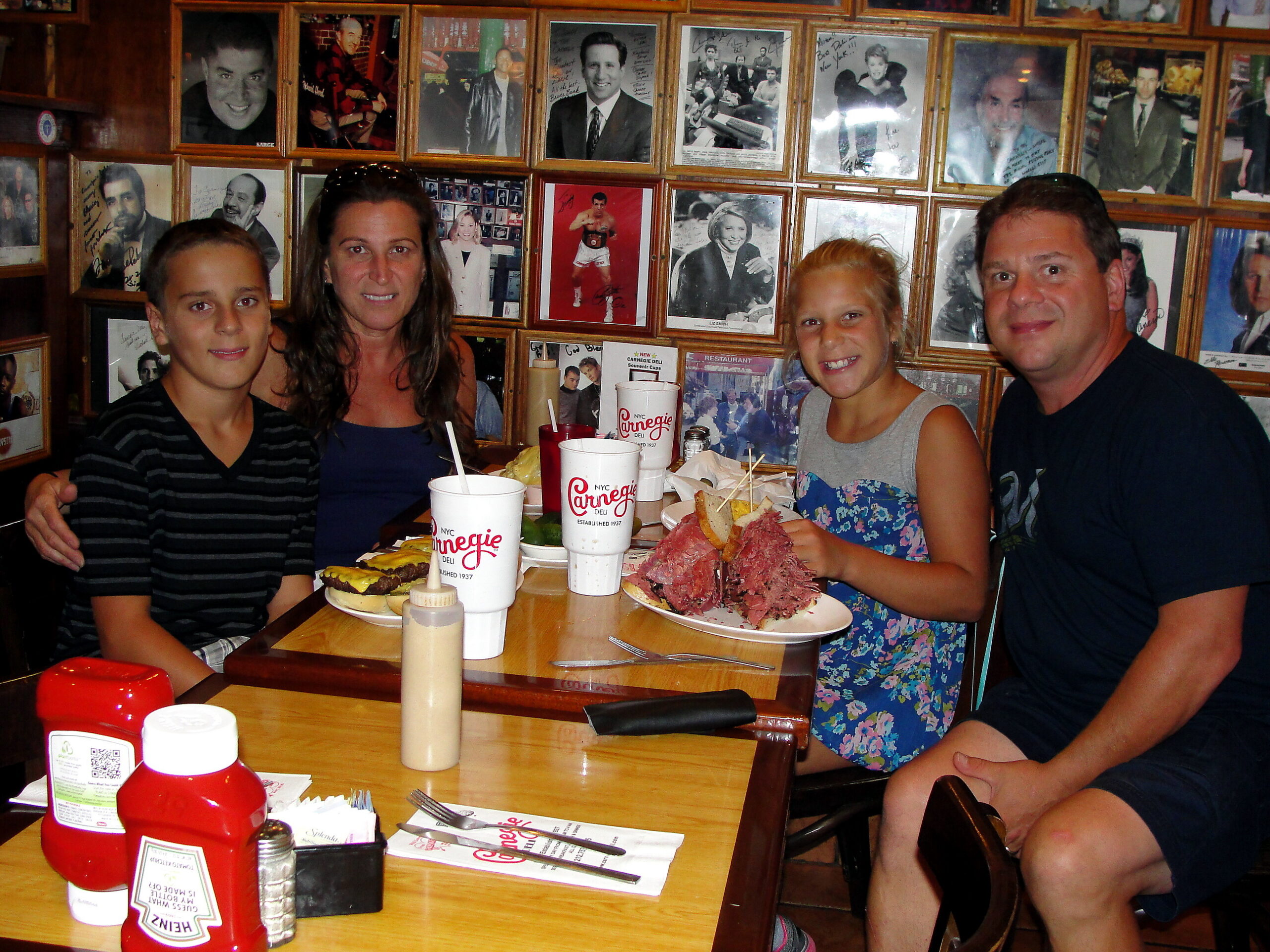 Group of four people smiling at a restaurant table with plates of food and drinks, in front of a wall covered with framed autographs and photos.