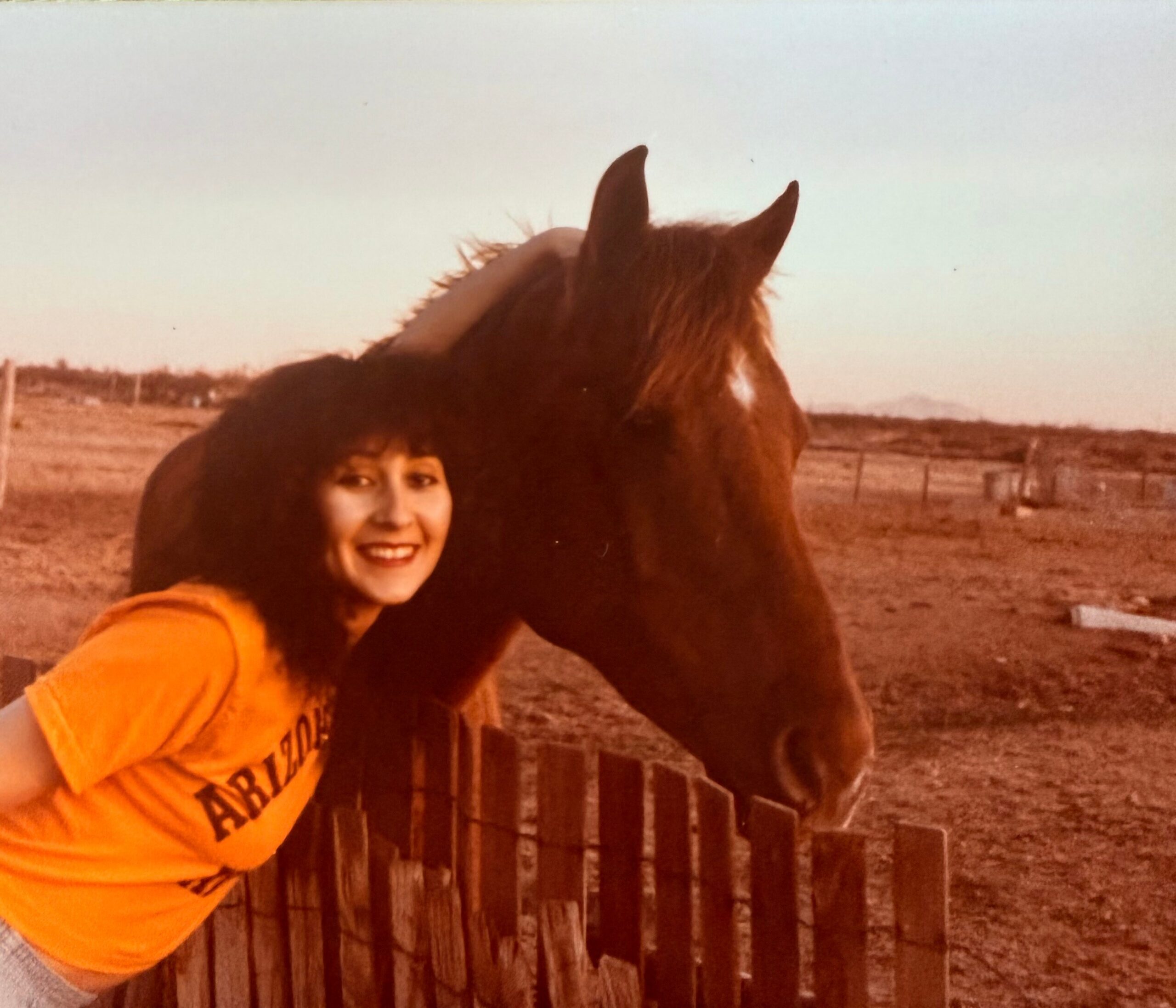 Person wearing an orange shirt stands beside a brown horse by a wooden fence in a rural setting, smiling at the camera.