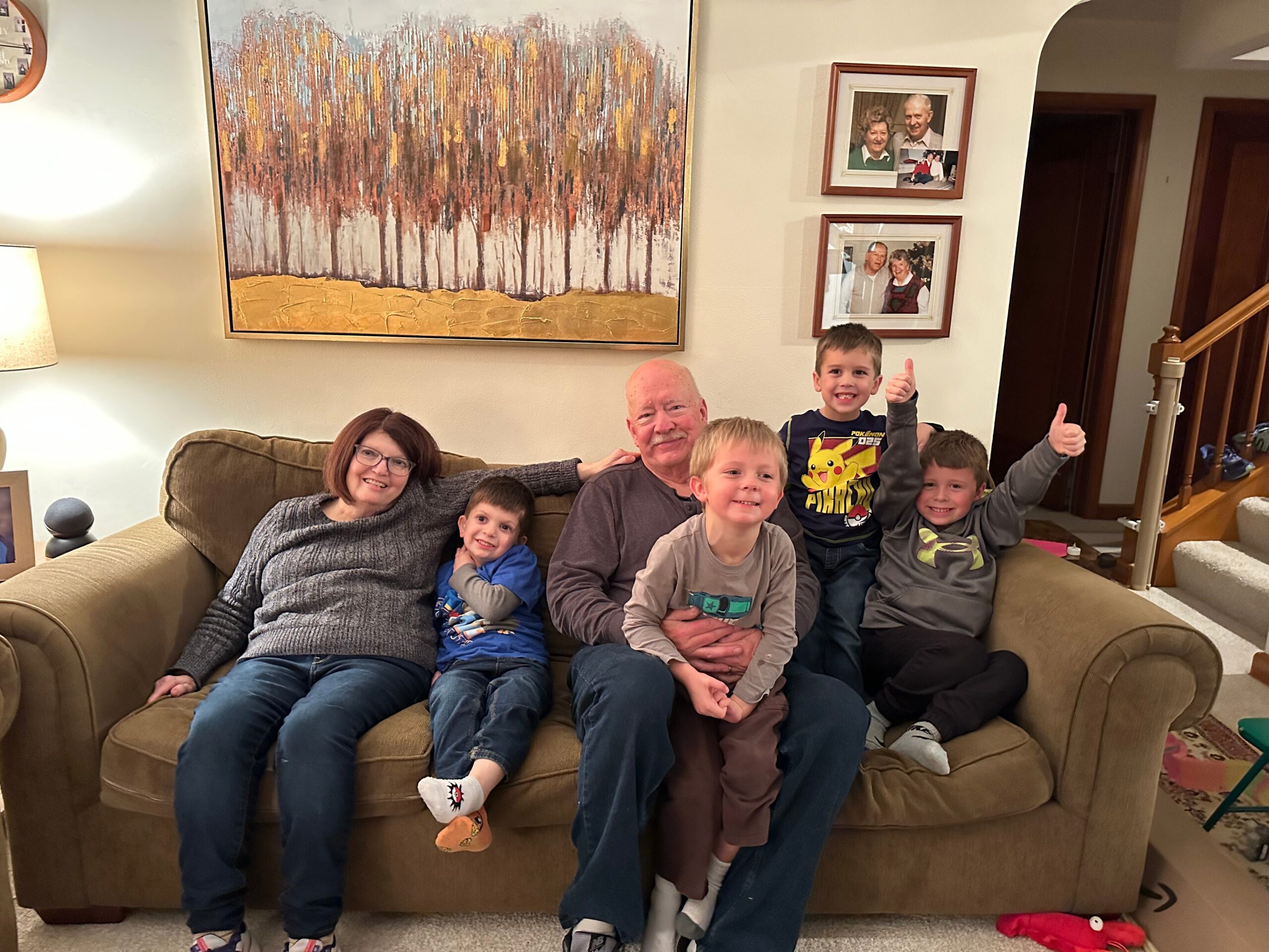 Family group of adults and children sitting on a brown sofa in a living room, smiling at the camera with thumbs up.