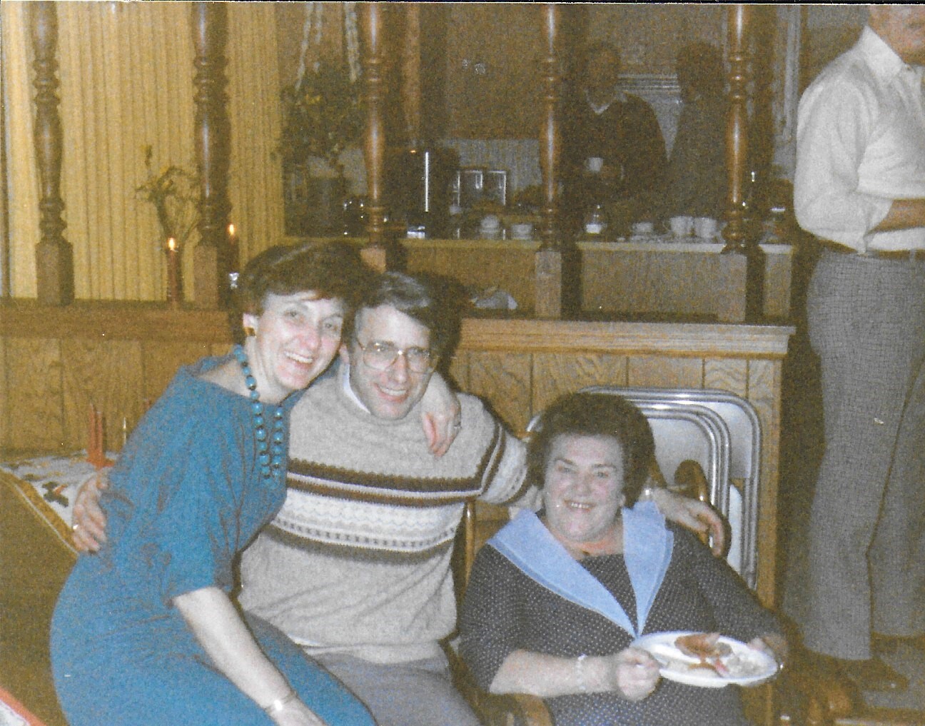 Three smiling friends pose together in a cozy, wood-paneled room at a casual gathering; one seated with a plate of food.