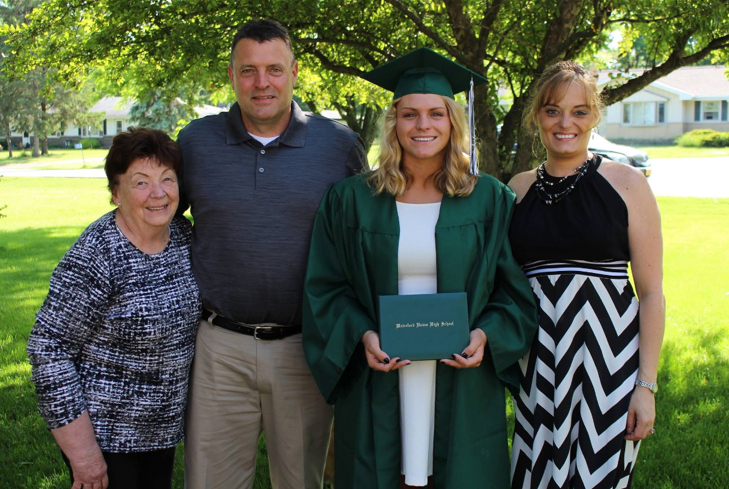 Graduate in green cap and gown holding diploma, posing with two relatives outdoors on a sunny day.