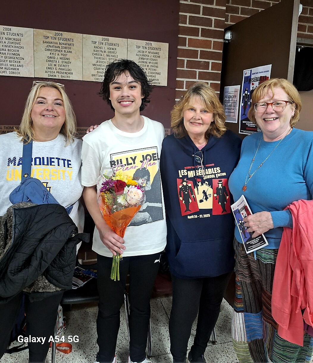 Four people smile for a group photo indoors; the young man in the center holds a bouquet of flowers.