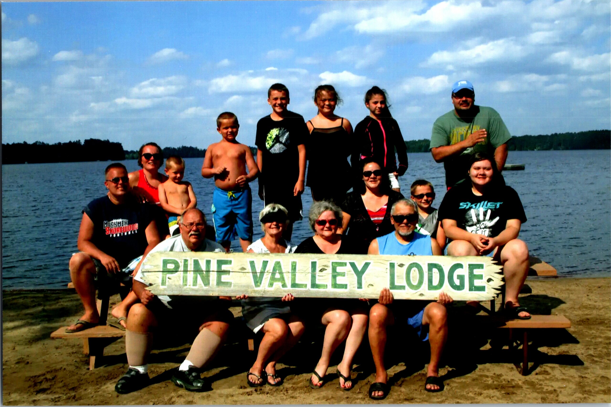Group of people posing on a wooden bench by a lake, holding a sign that reads 'PINE VALLEY LODGE' on a sunny beach.