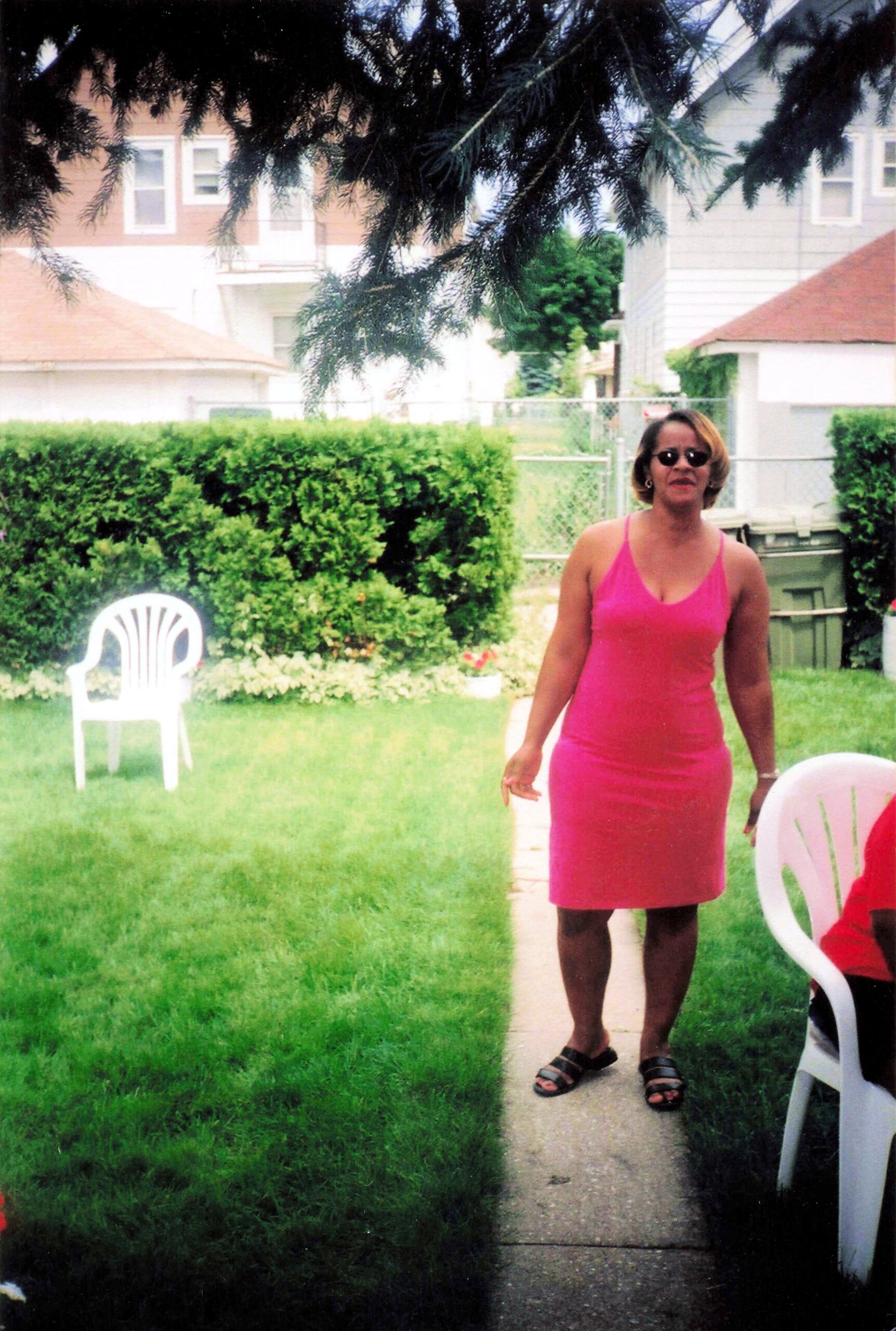 Woman in a bright pink dress and sunglasses standing on a concrete path in a suburban backyard, with hedges and plastic chairs nearby.