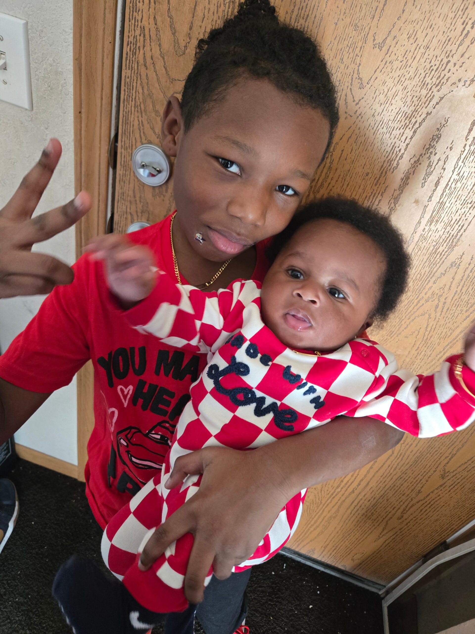 Two children hug in a doorway: an older boy in a red shirt making a peace sign while holding a younger child in a red-and-white checkered outfit.