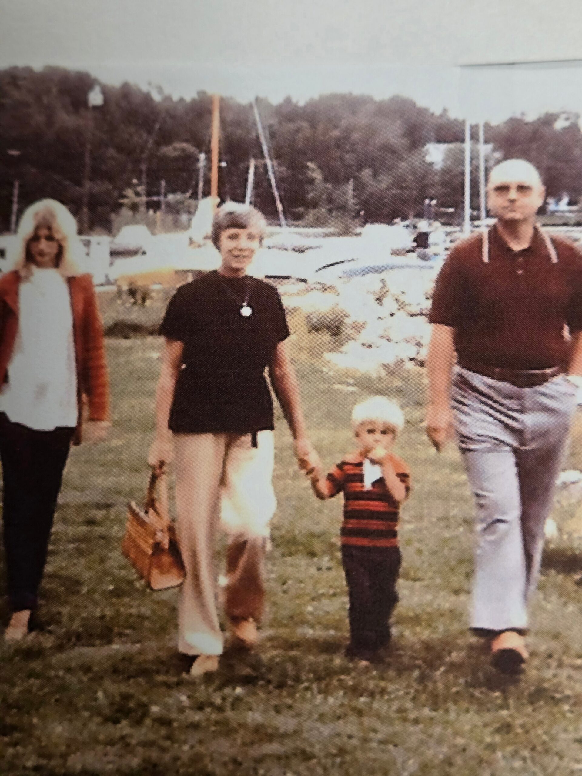 A family of four (two women, a man, and a small child) walks on grass near a marina with boats in the background, holding a child's hand and a dog leash visible in the foreground.